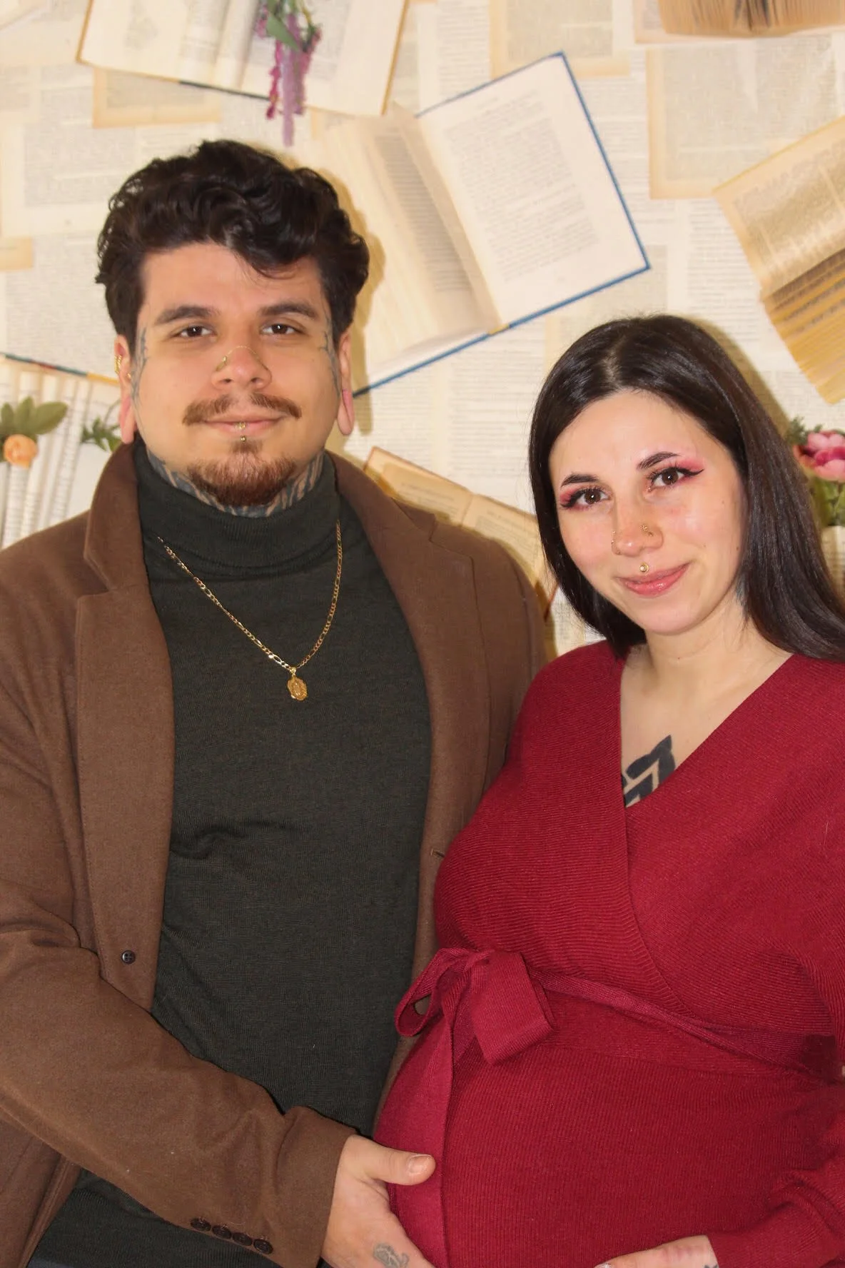 A couple, a man and a pregnant woman, standing together indoors in front of a decorative wall with hanging open books and flowers. They are smiling and posing for the photo.