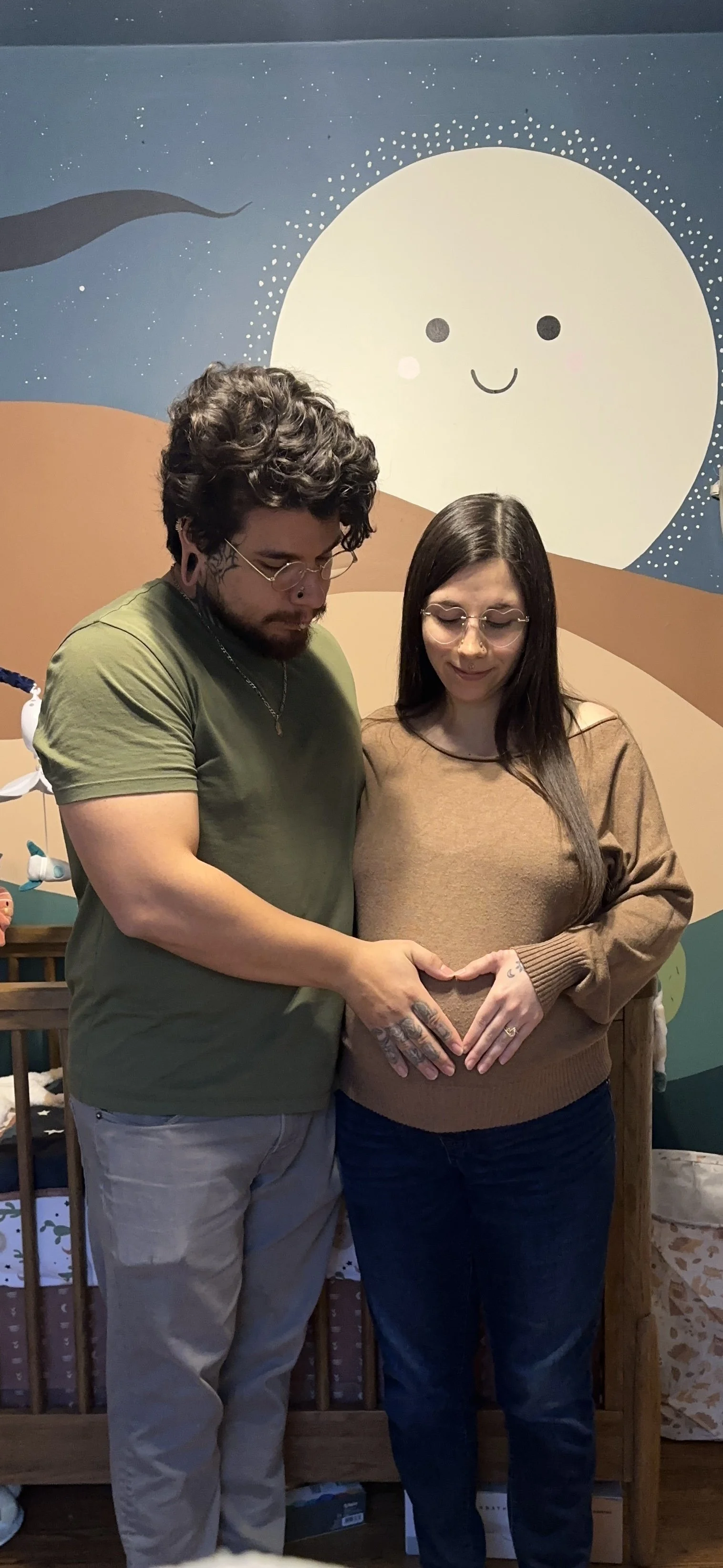 A man and woman standing close together, touching their hands on the woman's pregnant belly, forming a heart shape with their hands inside a children's room with a colorful mural of a smiling moon in the background.