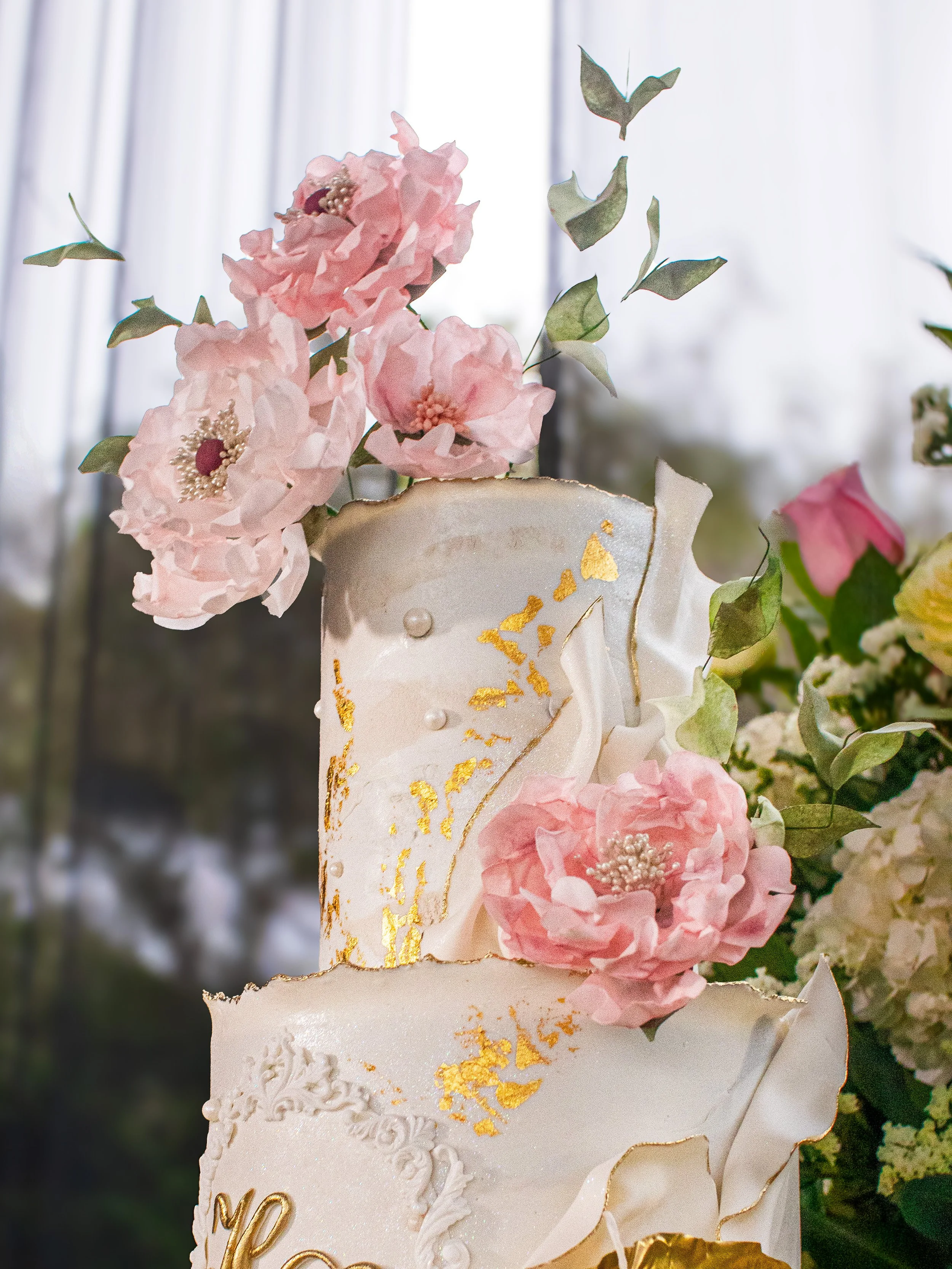 Close-up of a multi-tiered wedding cake decorated with pink flowers, green leaves, and gold accents.