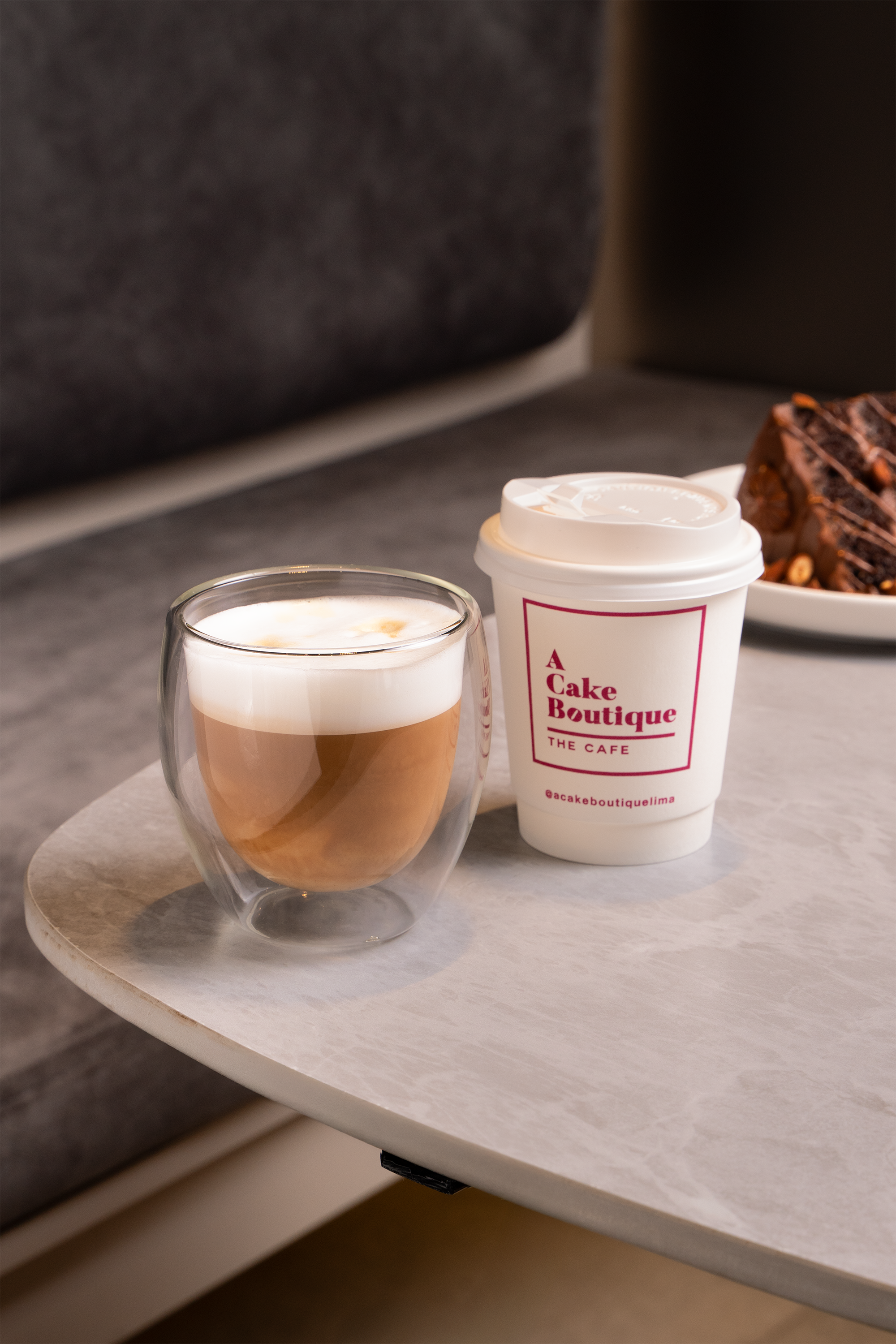 A cup of latte with foam art, a disposable coffee cup labeled "A Cake Boutique The Cafe," and a plate of chocolate cake on a marble table.