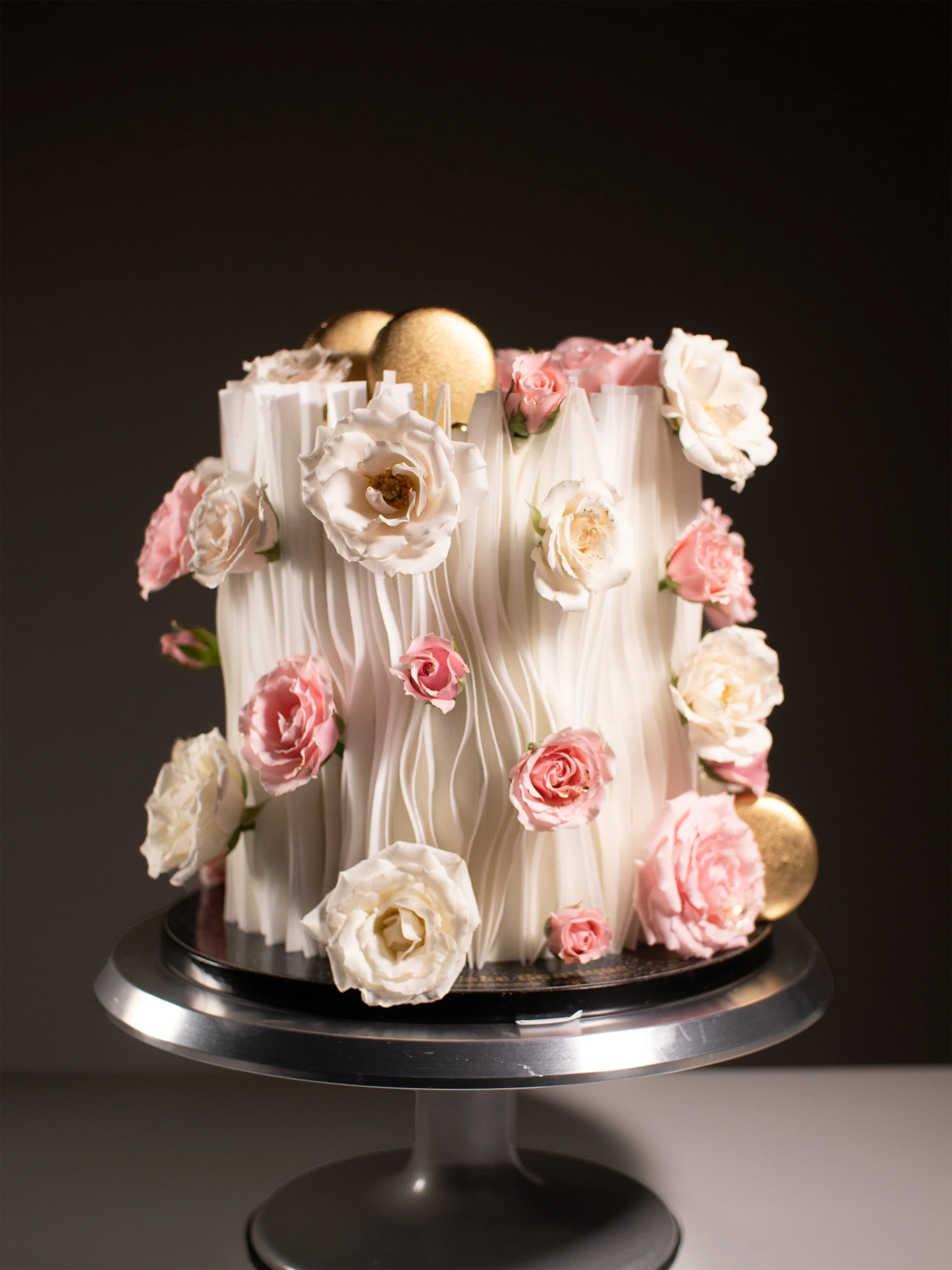 A tall, cylindrical cake decorated with pink and white roses, white chocolate shavings, and gold spheres on a metallic cake stand.