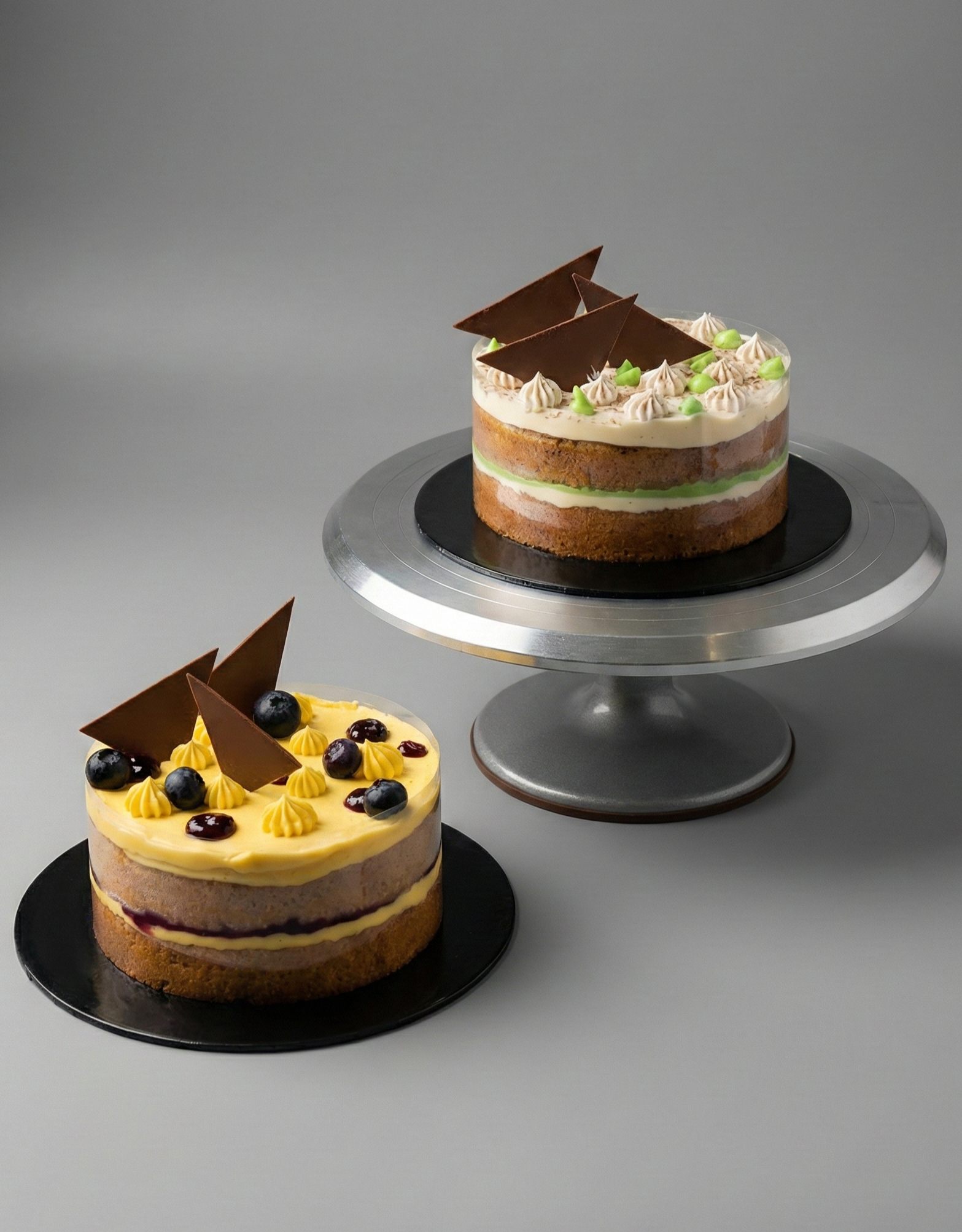 Two layered cakes decorated with chocolate triangles, fruit, and frosting on black and silver cake stands.