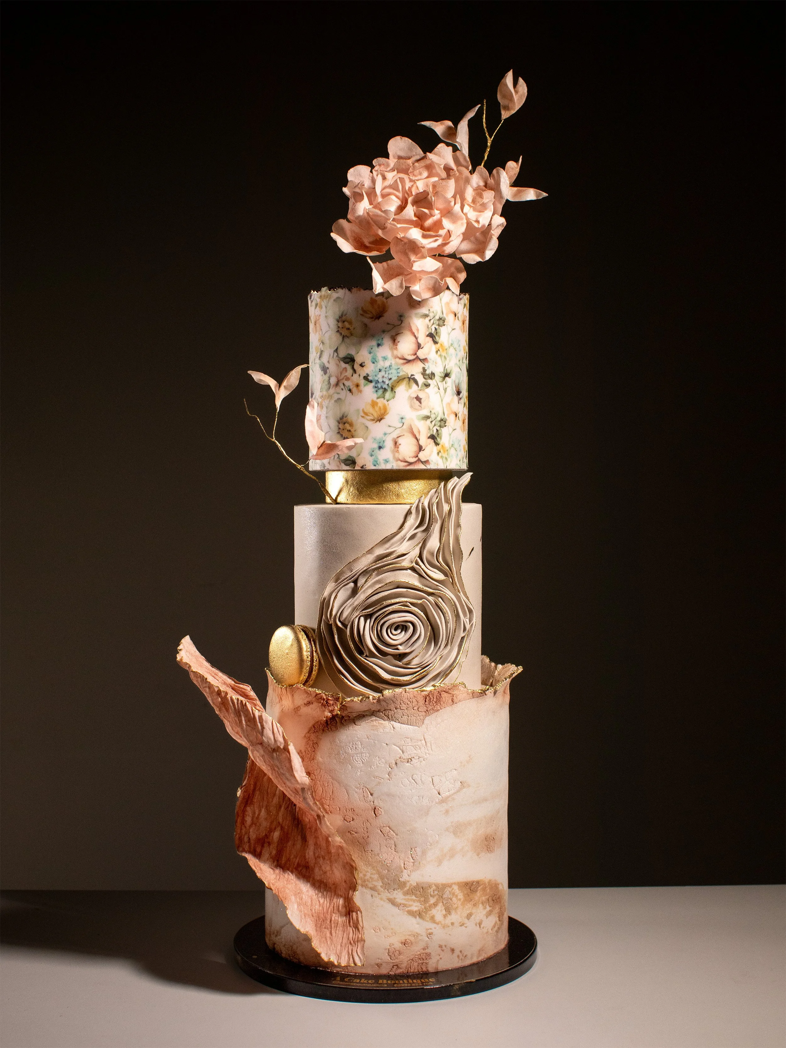 A three-tiered cake with floral and artistic decorations, including pink flowers, gold accents, and abstract designs, set against a dark background.