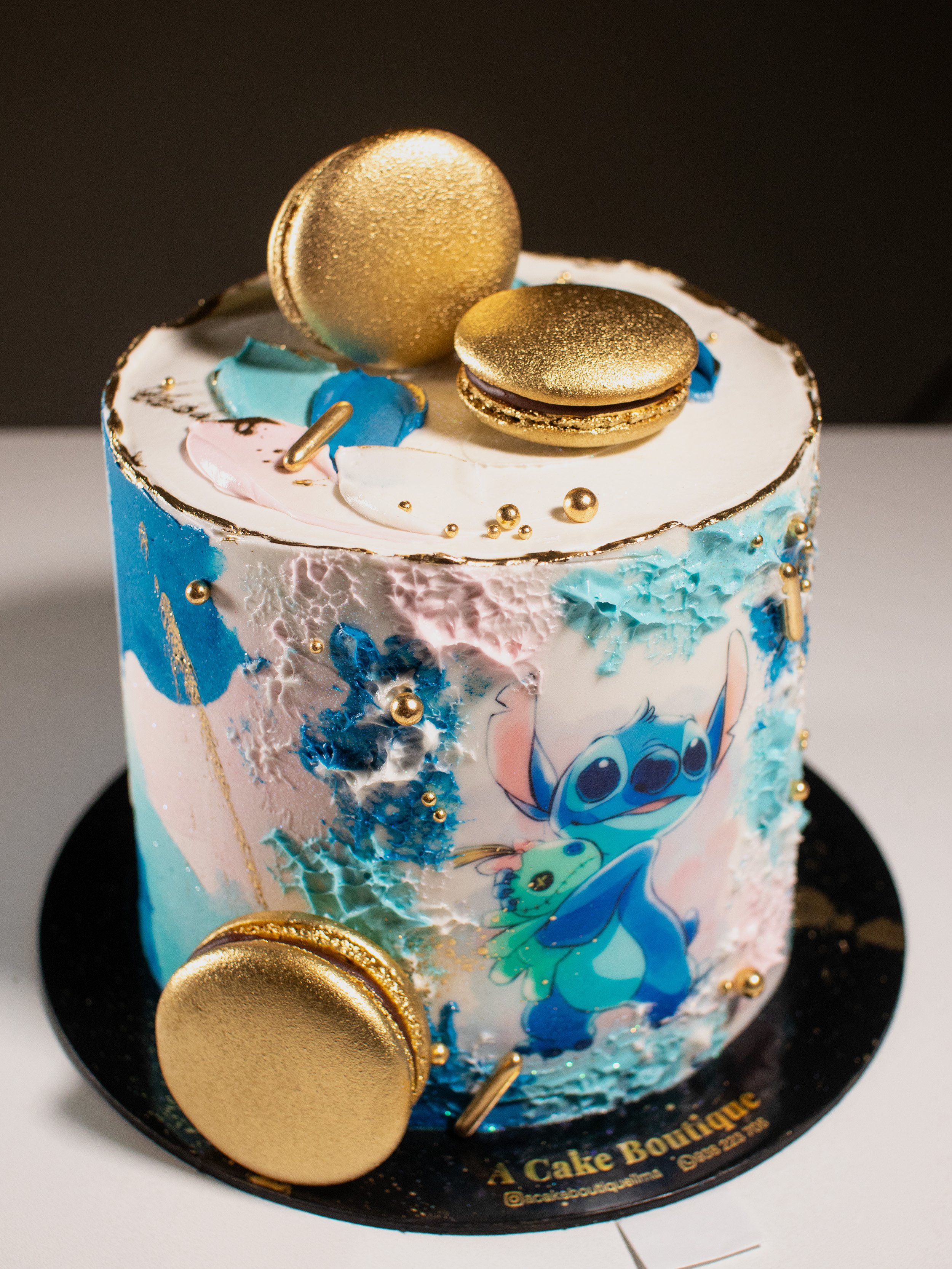 A decorated cake with a Stitch from Disney design, topped with gold macarons, colorful chocolate pieces, and gold accents.