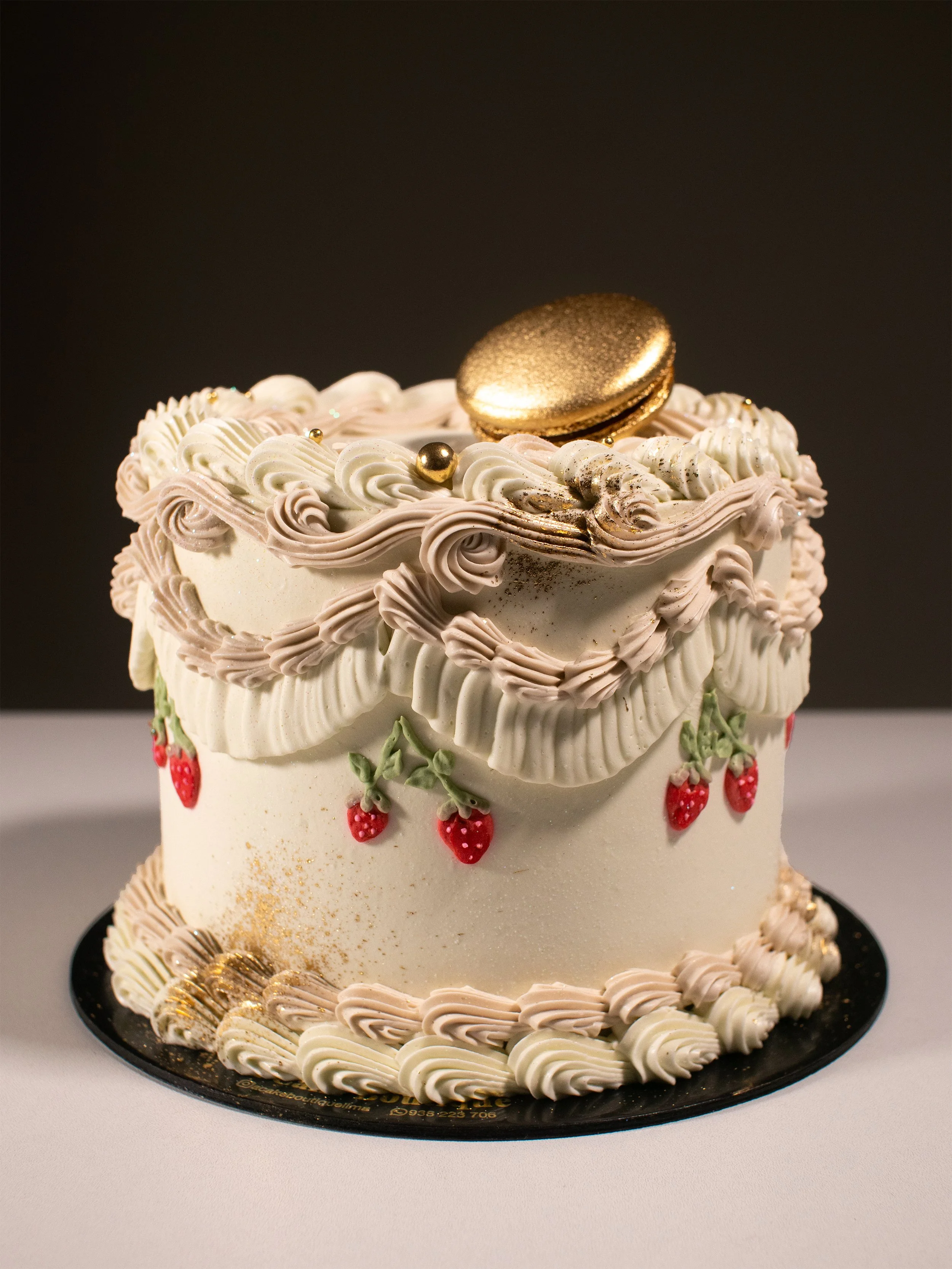 Decorated cake with white and pink frosting, strawberries, a gold macaron on top, and gold accents.