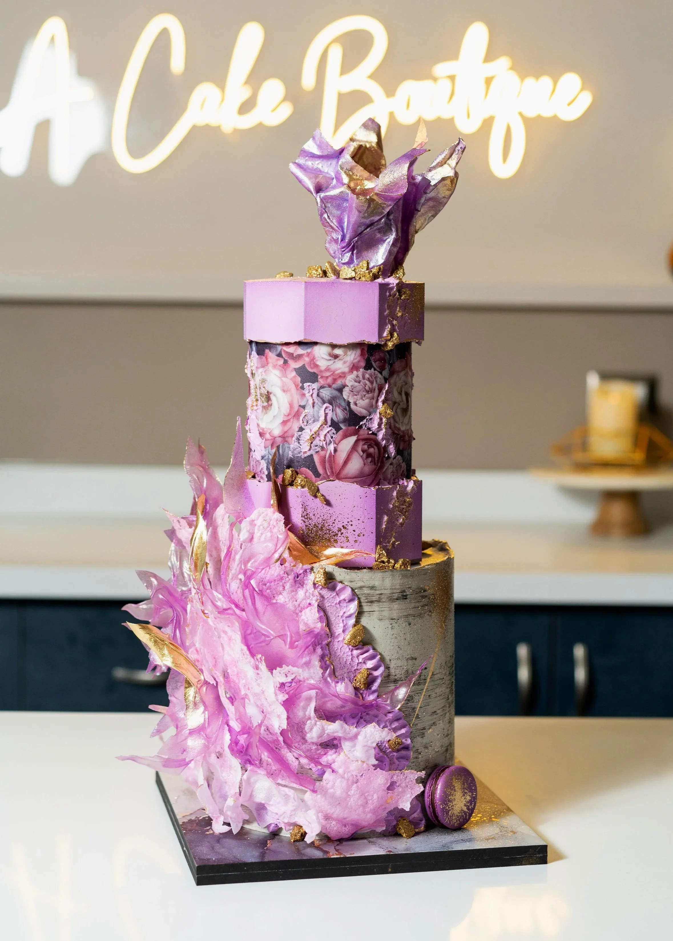 A tall, layered cake with a metallic silver bottom layer, pink and purple decorative blocks, floral patterns, and a purple and gold sculpted top decoration, on a black square base, with a neon sign that reads 'A Cake Boutique' in the background.