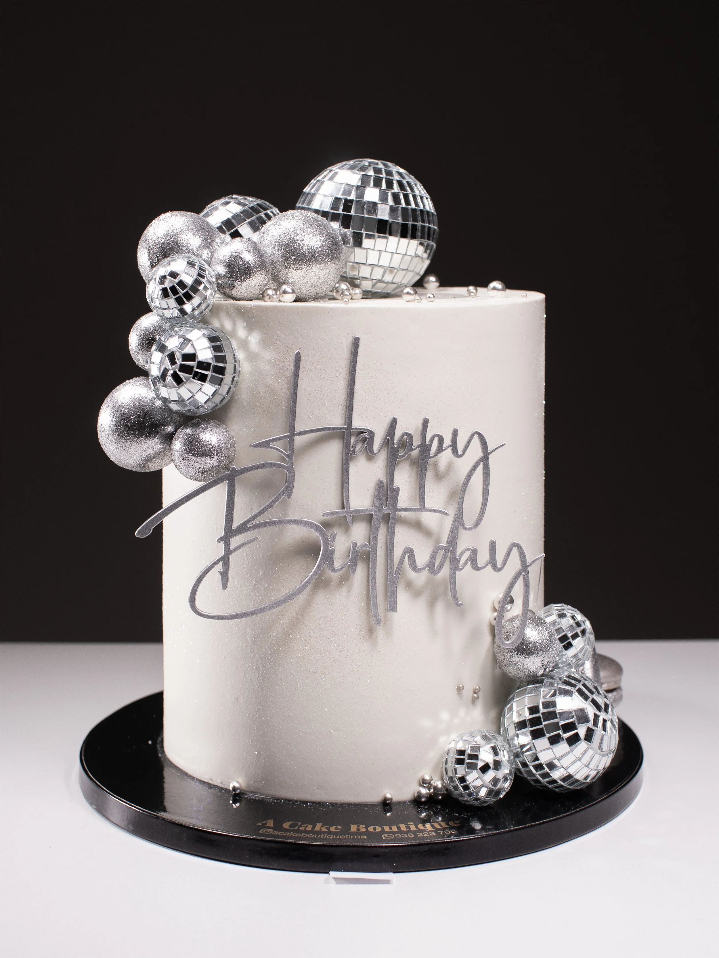 A white birthday cake decorated with silver disco balls and a "Happy Birthday" topper, on a black cake board.