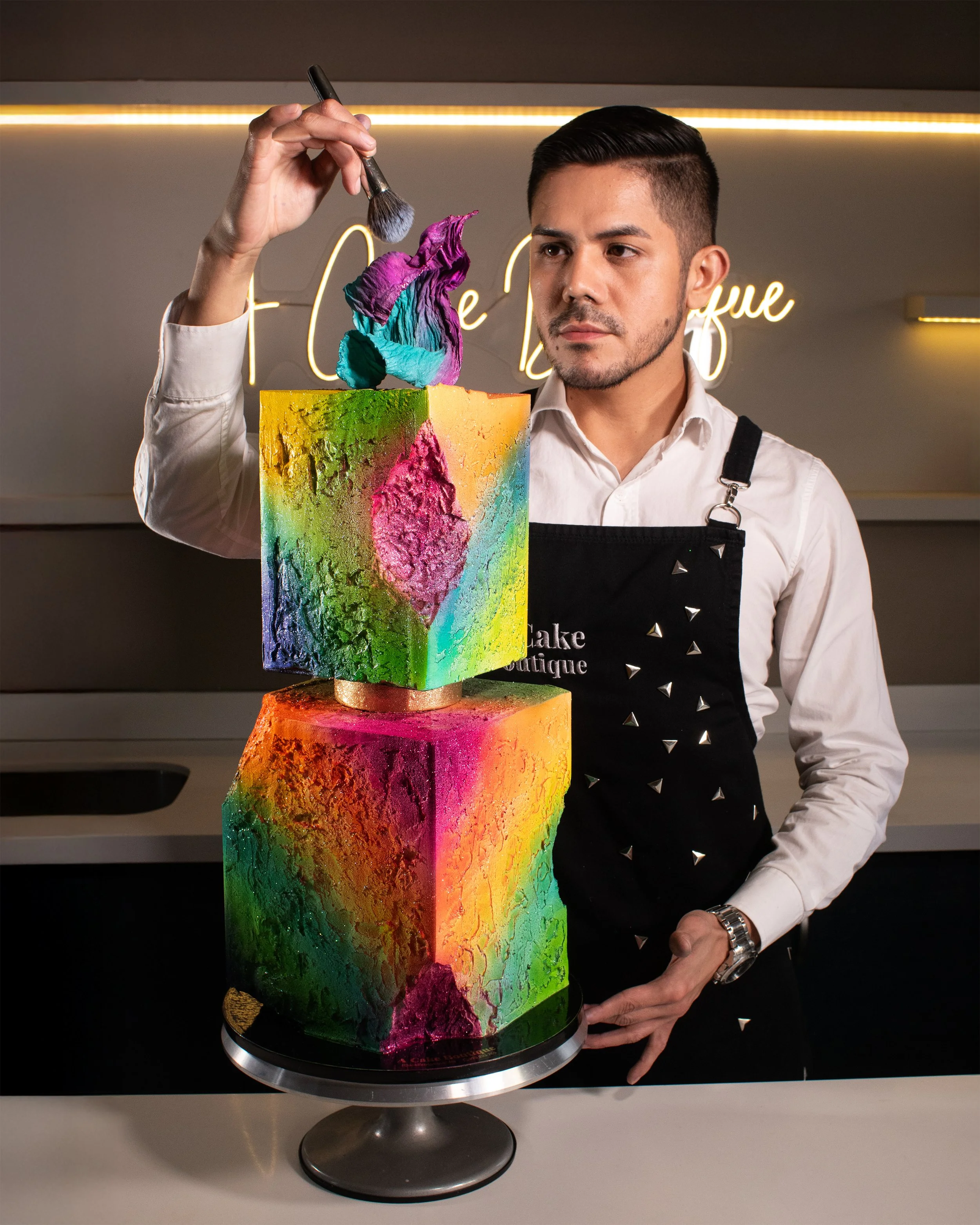 A chef is decorating a colorful, abstract cake with a paintbrush in a bakery or cake shop setting.