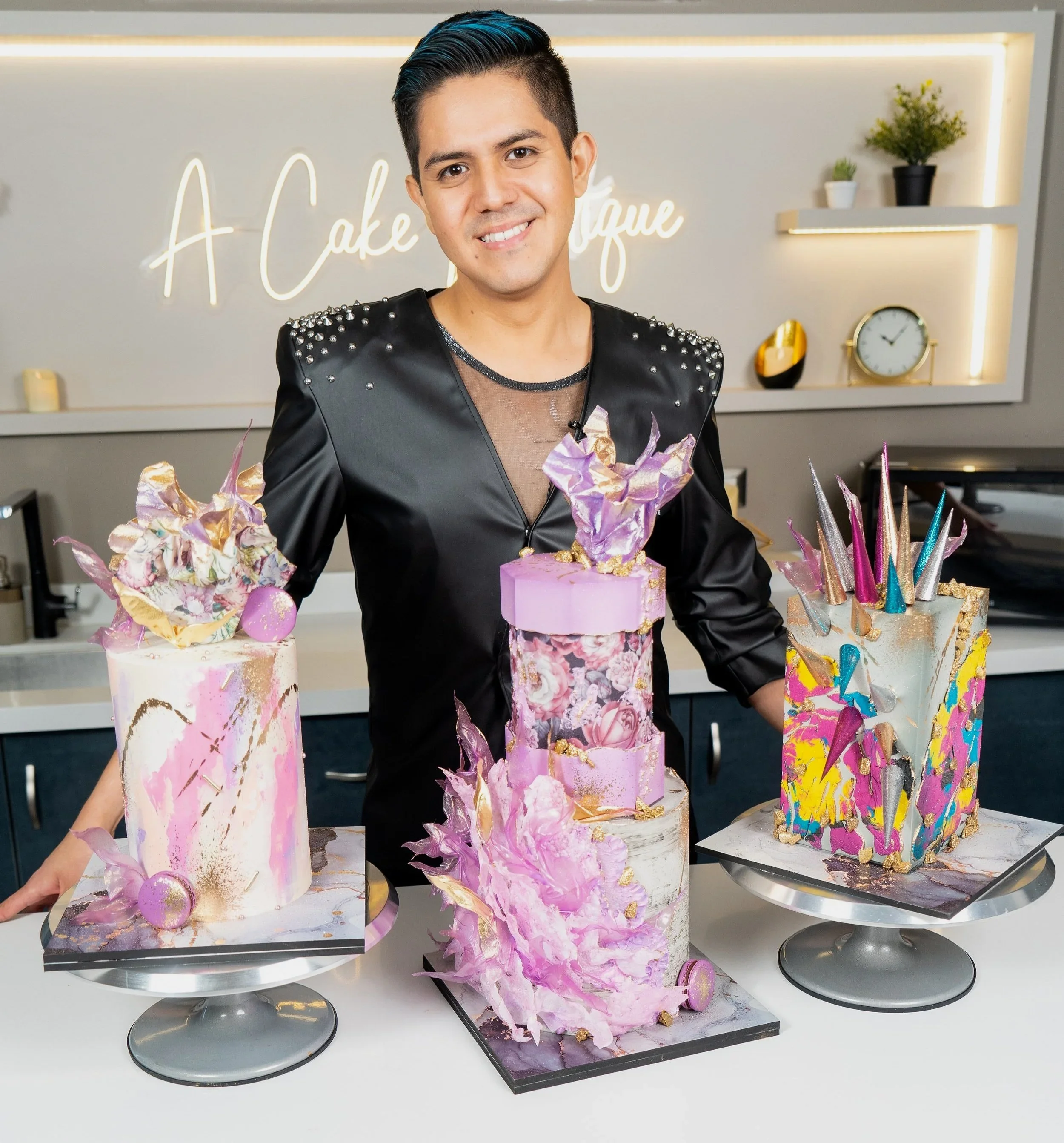 A man in a black satin jacket with pearl embellishments stands behind three colorful, artistic cakes on display. The background features a modern kitchen and a neon sign that reads 'A Cake Boutique'.