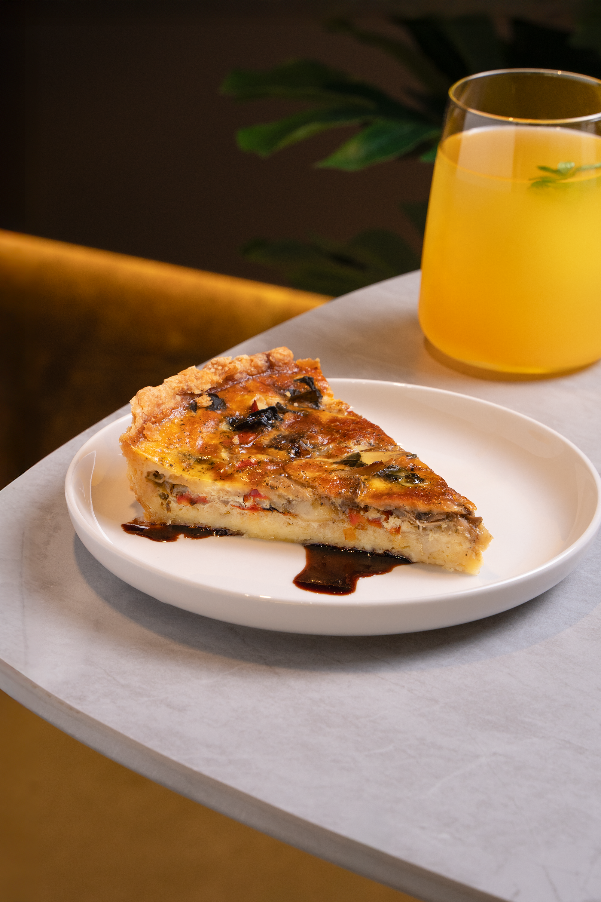 A slice of vegetable quiche on a white plate with a side of dark sauce, next to a glass of yellow-orange beverage, on a light-colored tabletop.