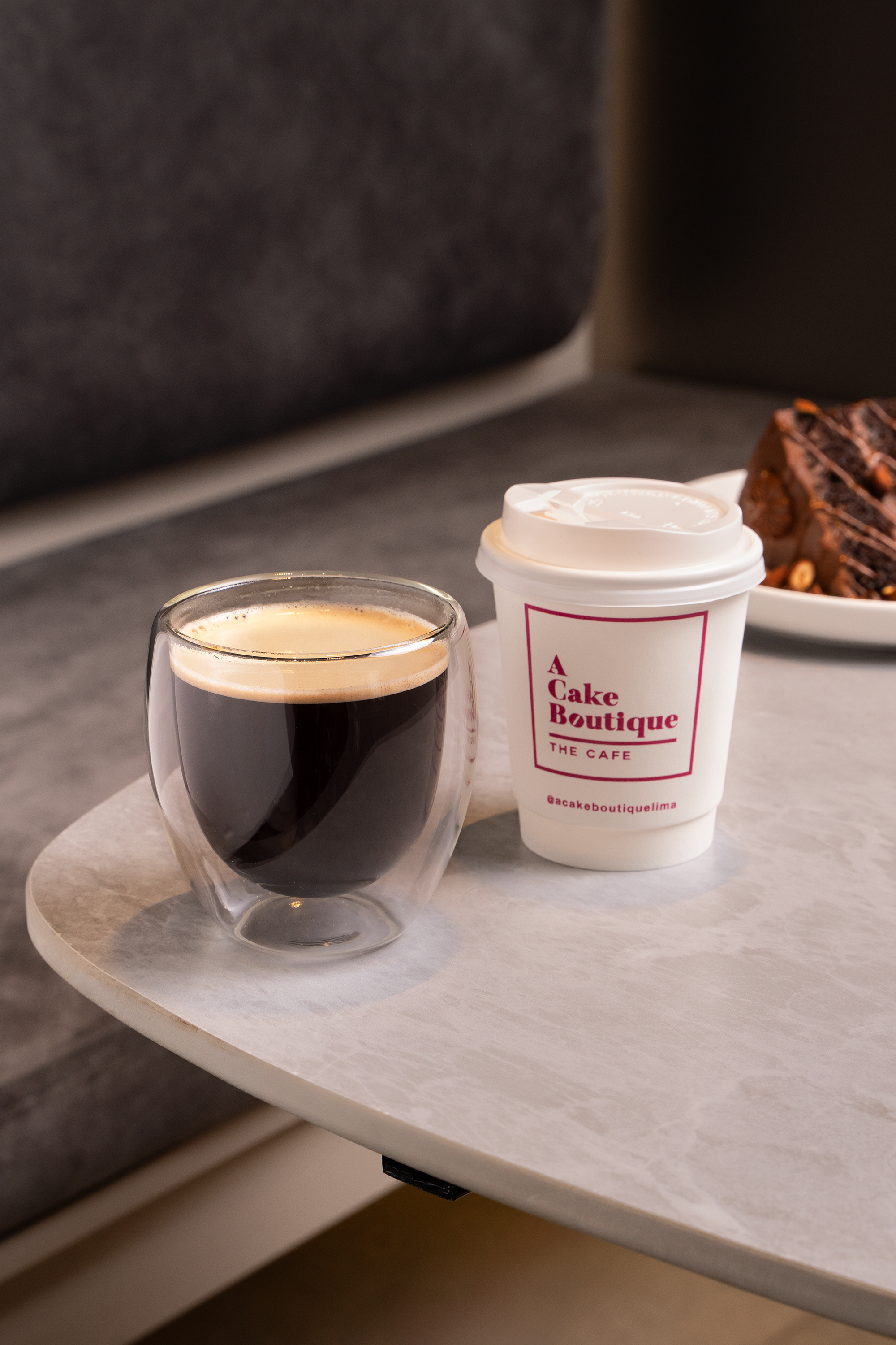 A coffee cup on a table with a logo reading 'A Cake Boutique', a glass cup of black coffee, and a plate with a chocolate cake in the background.
