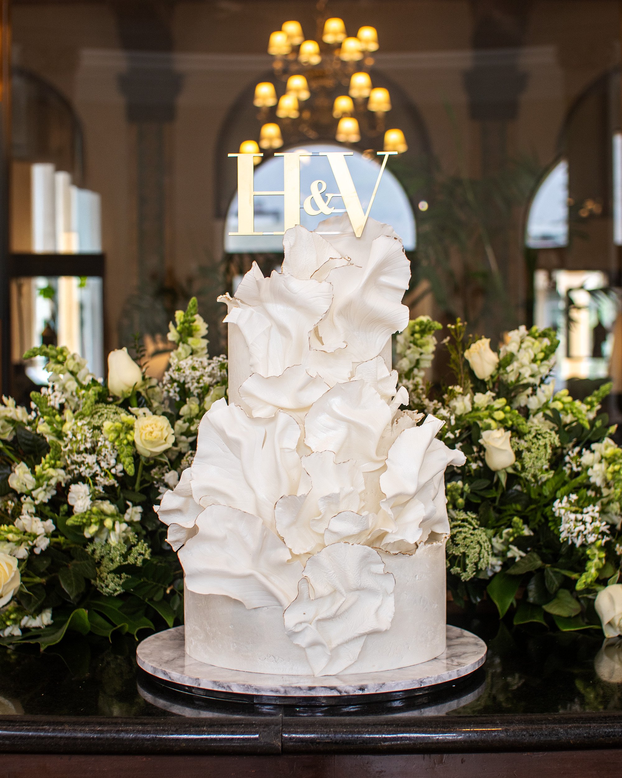 White wedding cake with ruffled icing and floral decorations, topped with a gold monogram "H & V" sign, surrounded by white flowers and greenery.