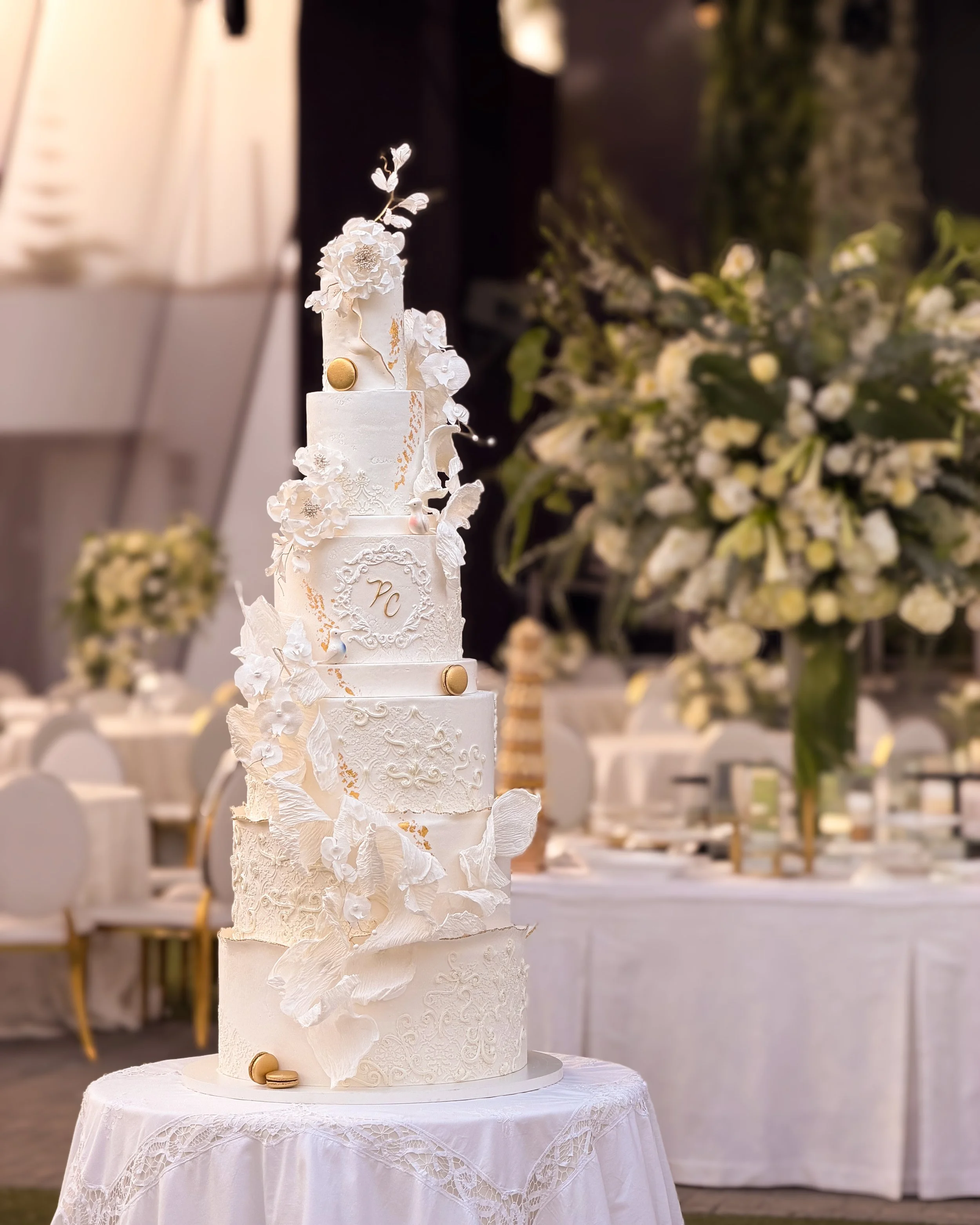 A tall, white wedding cake decorated with white flowers, gold accents, and intricate patterns, placed on a white lace-covered table at a wedding reception.