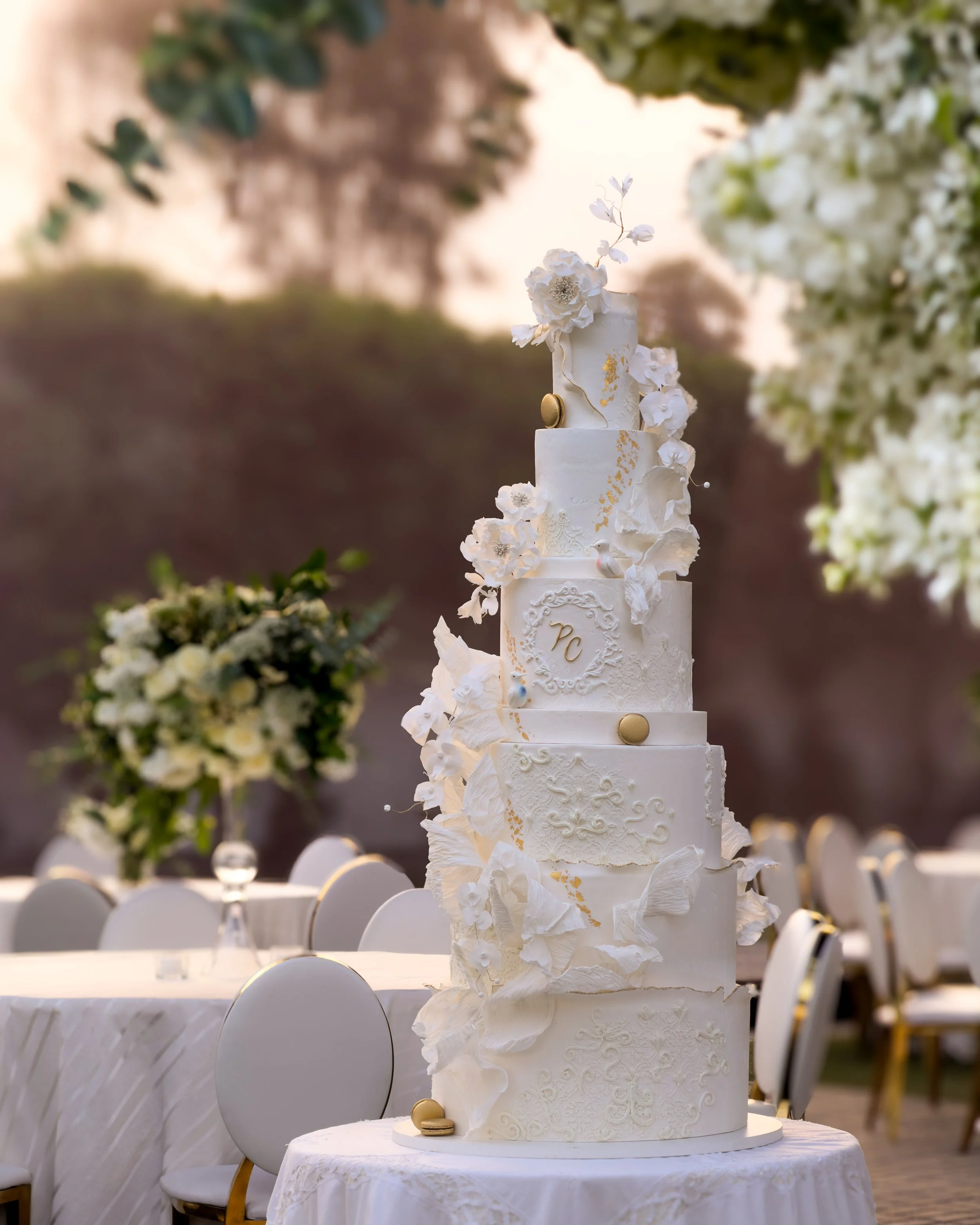 A tall, multi-tiered white wedding cake decorated with white flowers, gold accents, and monogram initials 'PC', set outdoors with tables and chairs in the background.