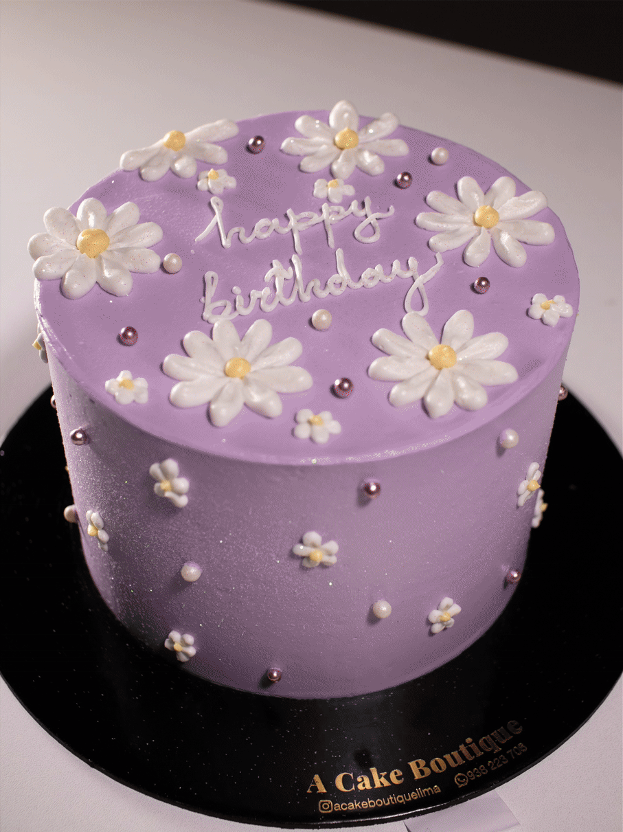 A purple birthday cake decorated with white flower-shaped icing and small edible pearls, with the words "happy birthday" written on top.