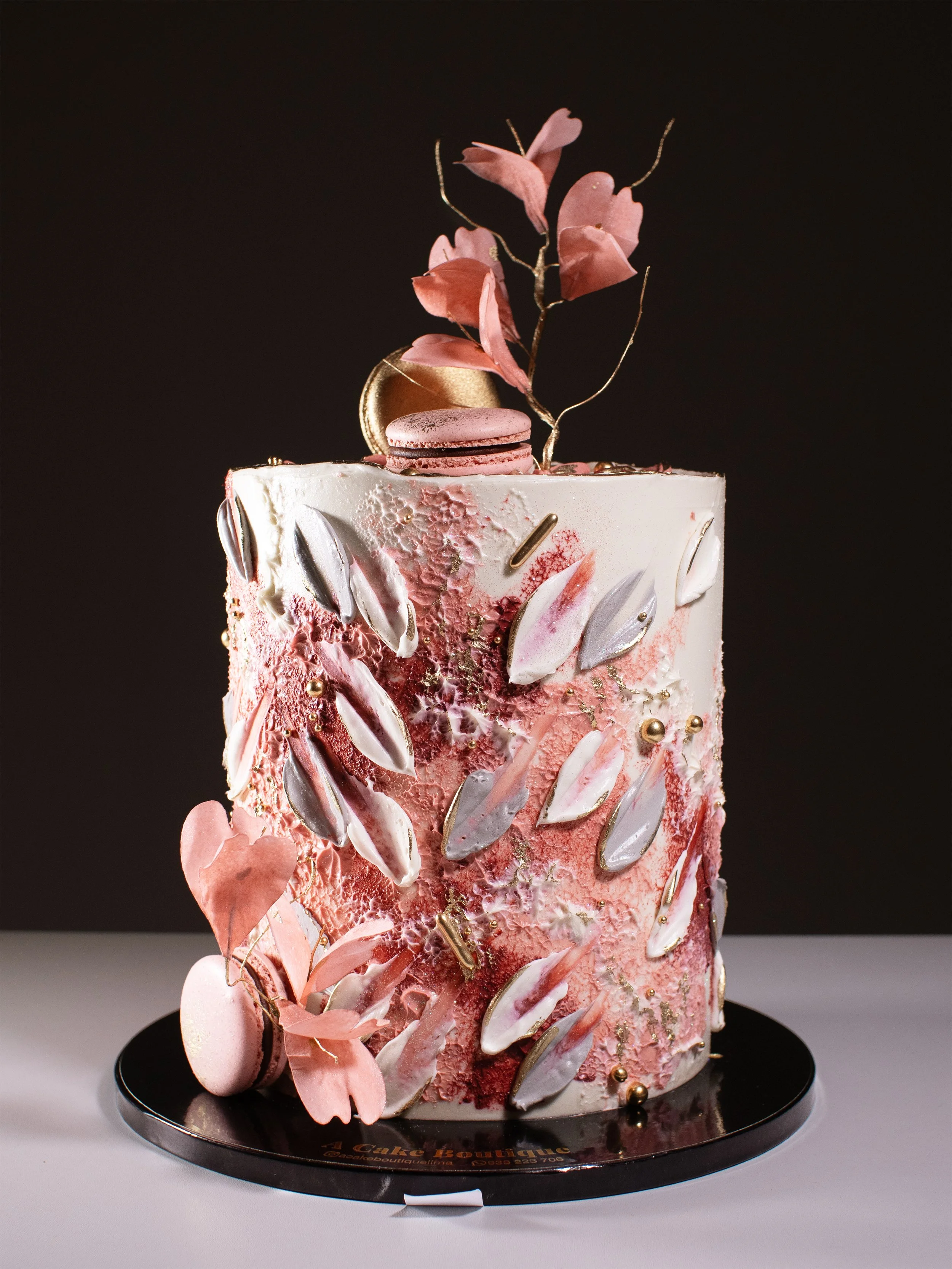 A tall, elaborately decorated pink and white cake with metallic accents, pink flowers, and macarons on top, displayed on a black circular base against a dark background.