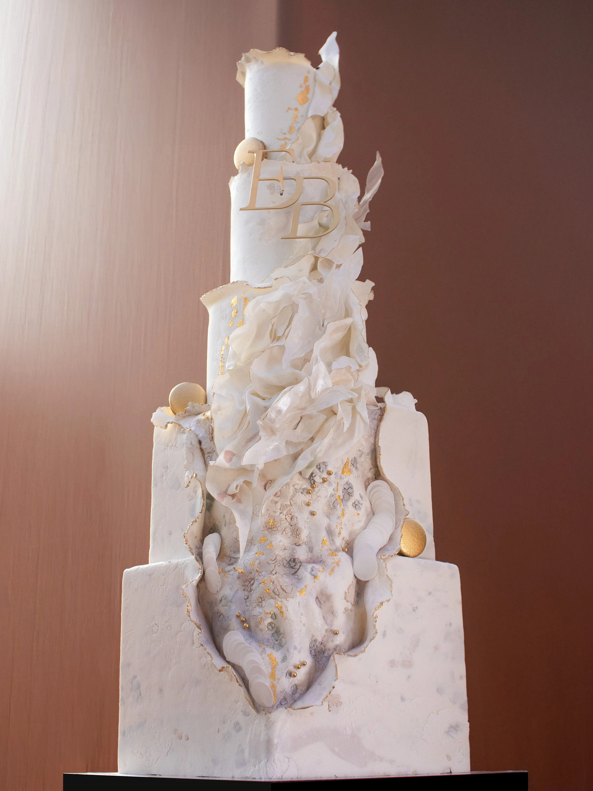 A tall, multi-tiered wedding cake decorated with white and gold accents, flowing white icing, and draped fabric elements.