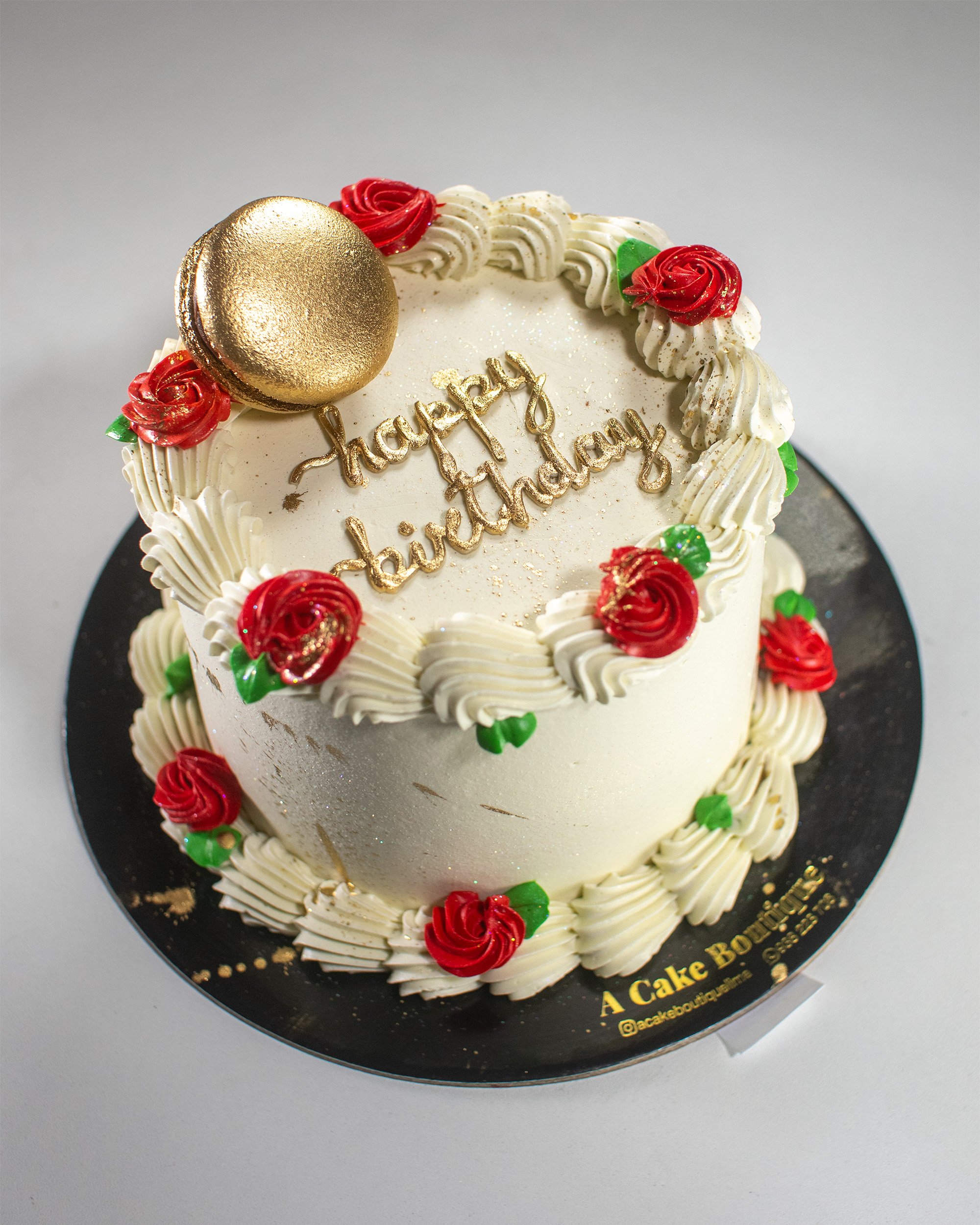 A white birthday cake decorated with red and green piping, gold lettering that says 'happy birthday,' and a gold spherical ornament on top, placed on a black cake board.
