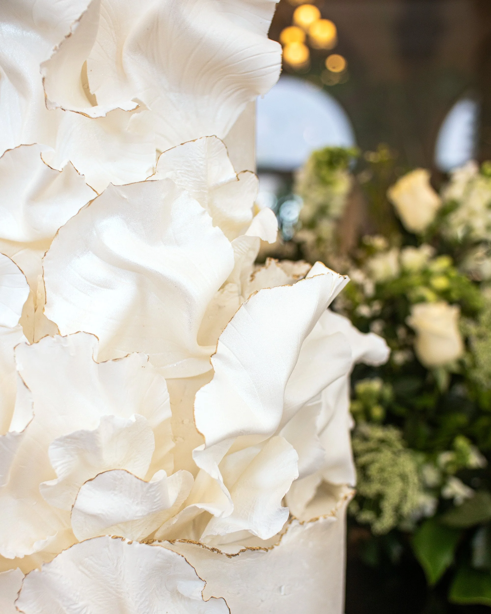 Close-up of white decorative flowers with gold edges, likely made of paper or fabric, with blurred background of floral arrangements.