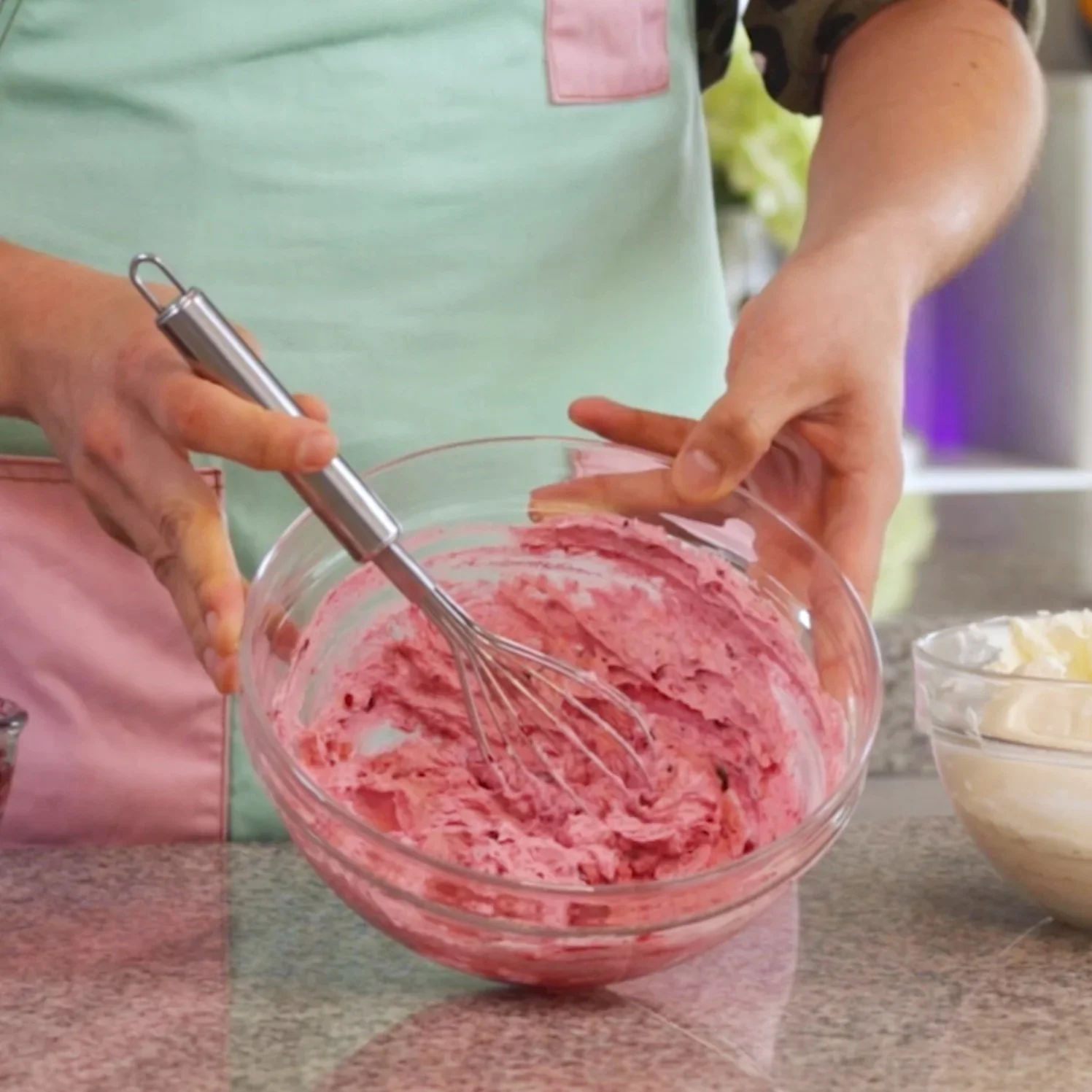 Person whisking pink and white striped frosting in a glass bowl on a kitchen countertop.