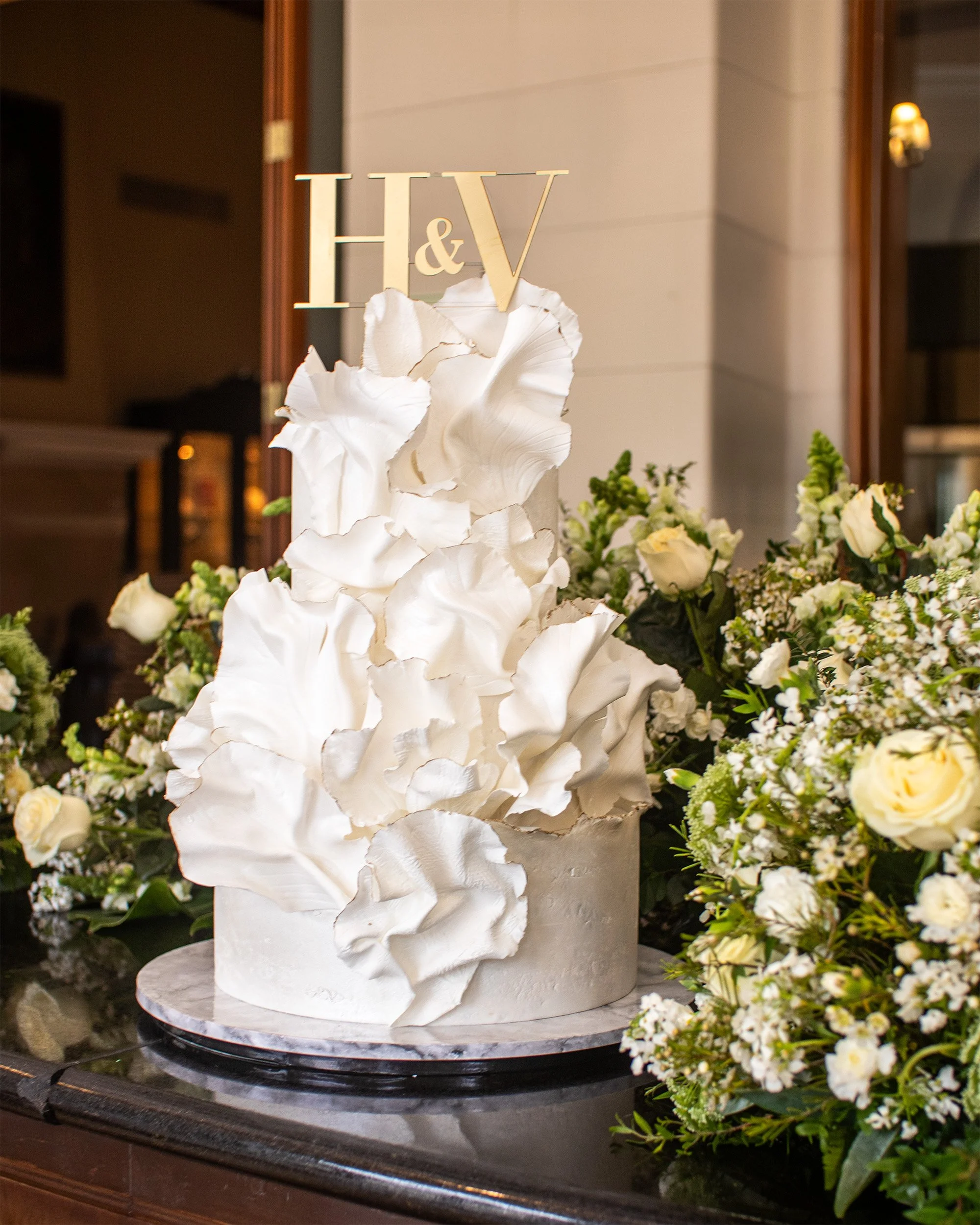 Elegant white wedding cake with large white sugar flower decorations, topped with gold initials 'H&V', surrounded by white and cream flowers including roses and small filler flowers.