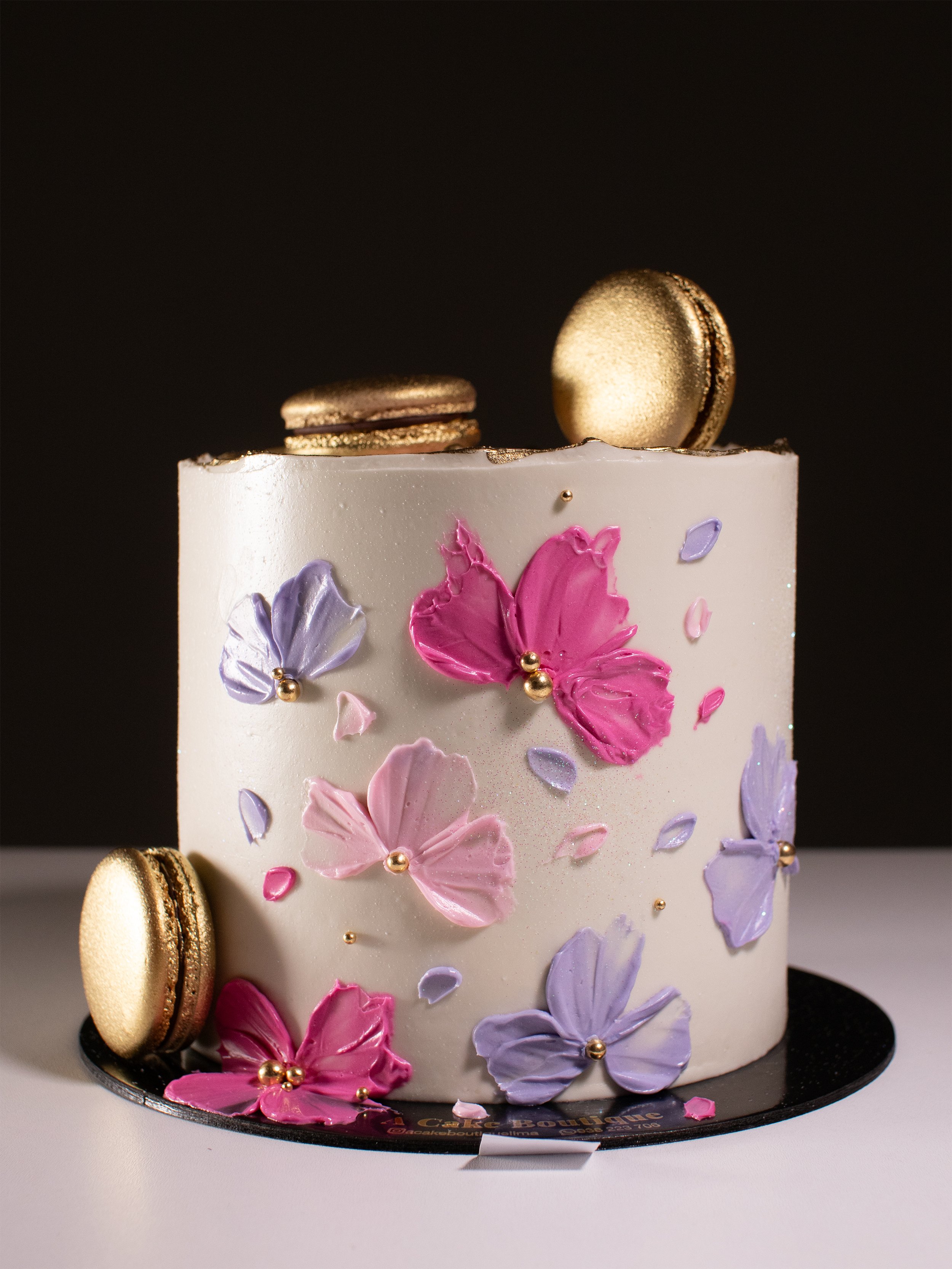 A white cake decorated with pink and purple butterfly-shaped flowers, topped with gold and brown macarons, on a black circular cake board against a dark background.
