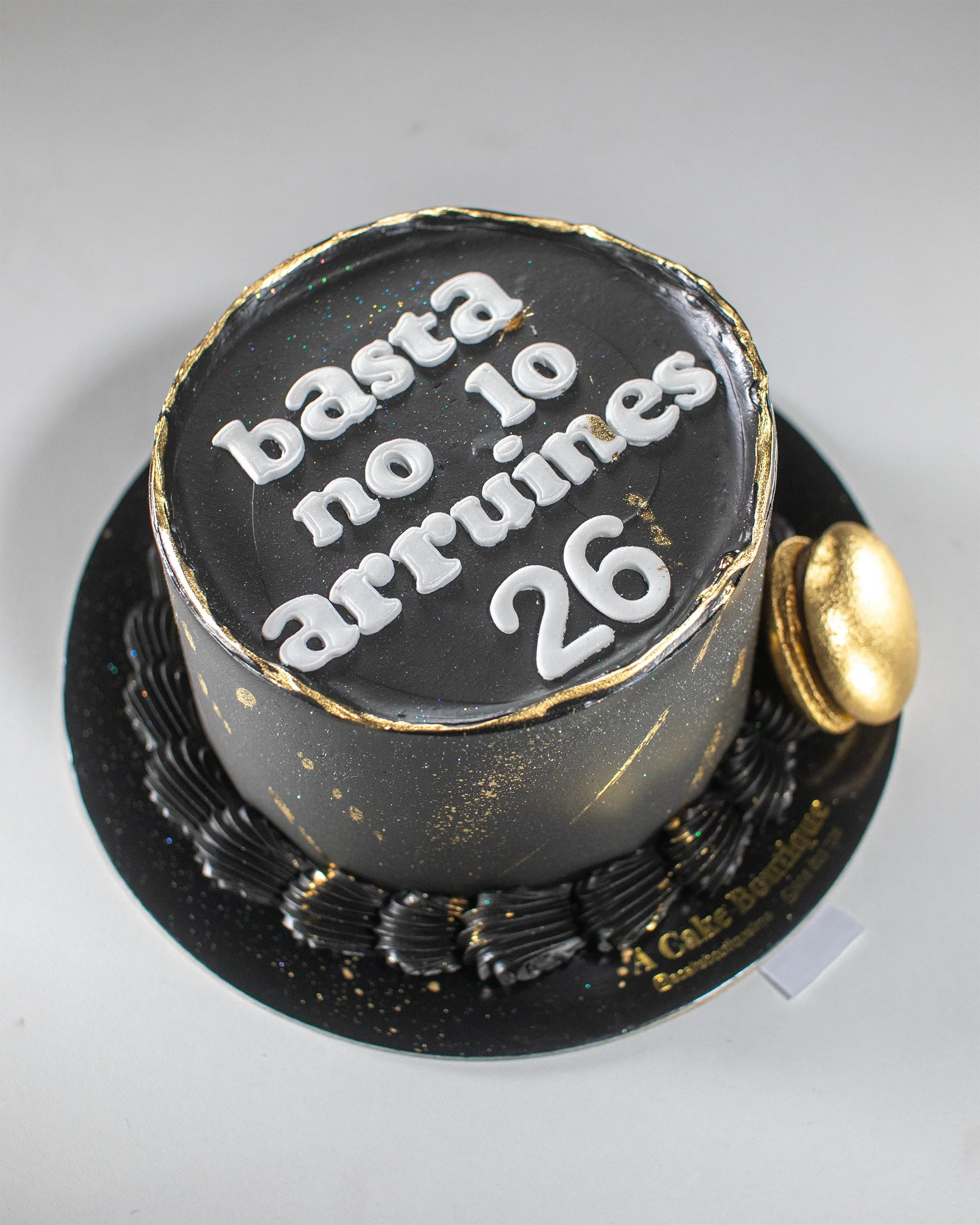 A round black cake with white letters reading 'basta no 10 airtrunes 26' on top, decorated with gold accents and a chocolate macaron, on a black cake board.