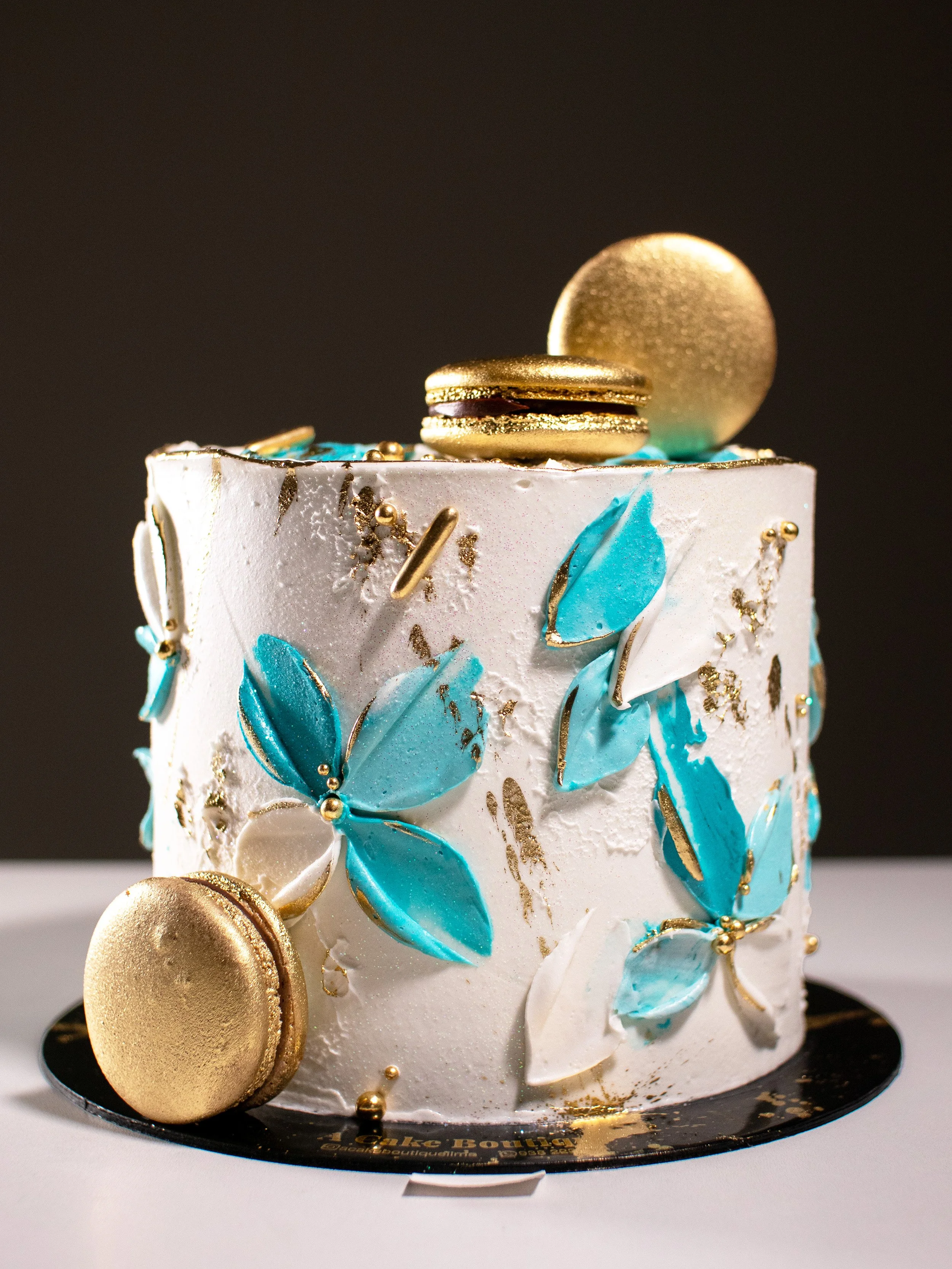 A decorated white cake with blue and white butterfly and leaf decorations, topped with a gold macaron and a gold coin, on a black cake board.