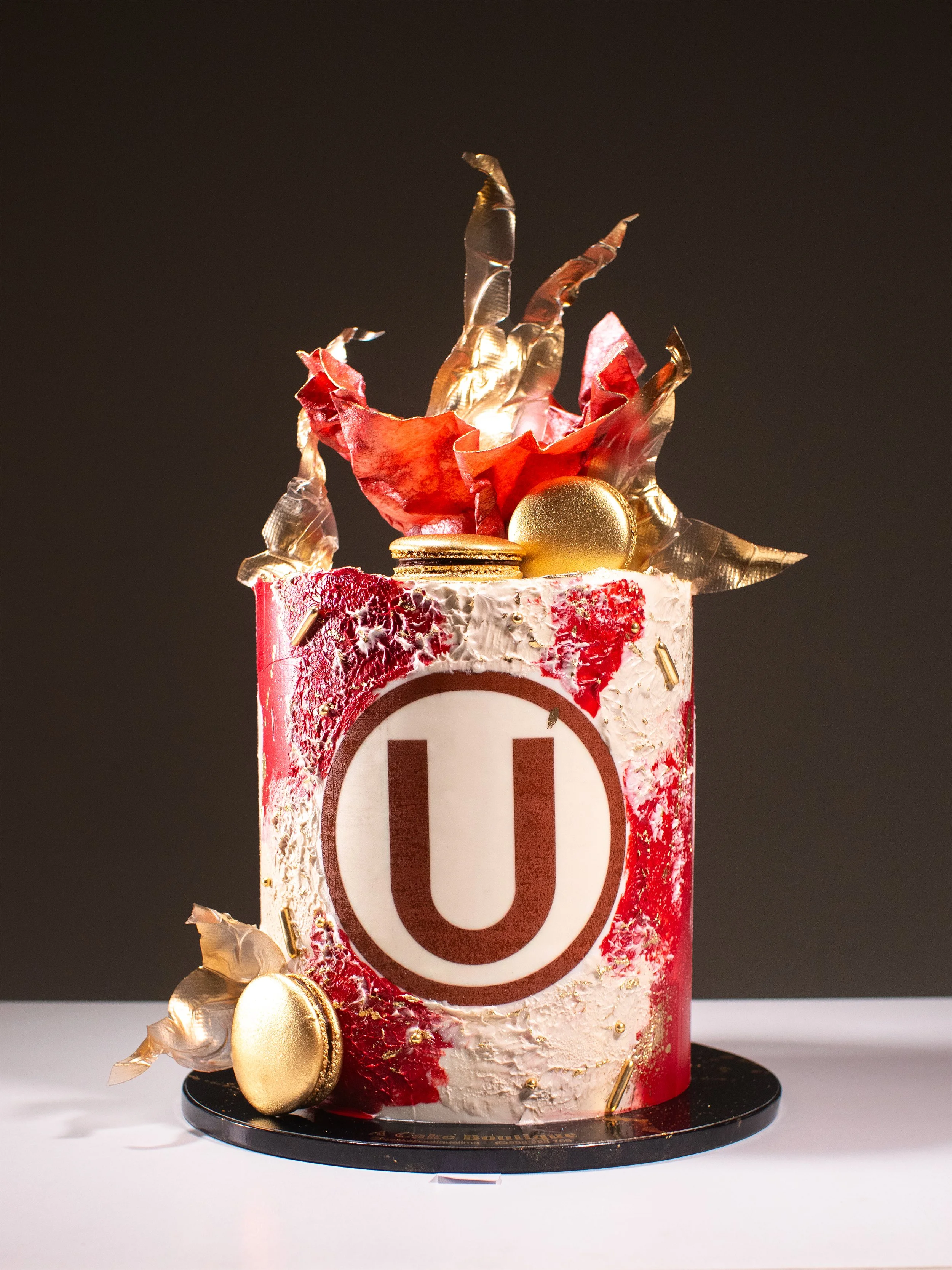 A decorated cake with a white and red textured icing, featuring a large U logo on the front. The cake is topped with gold and red decorative elements, including a large gold ball, gold macarons, and gold sprinkles, with artistic metallic sugar leaves