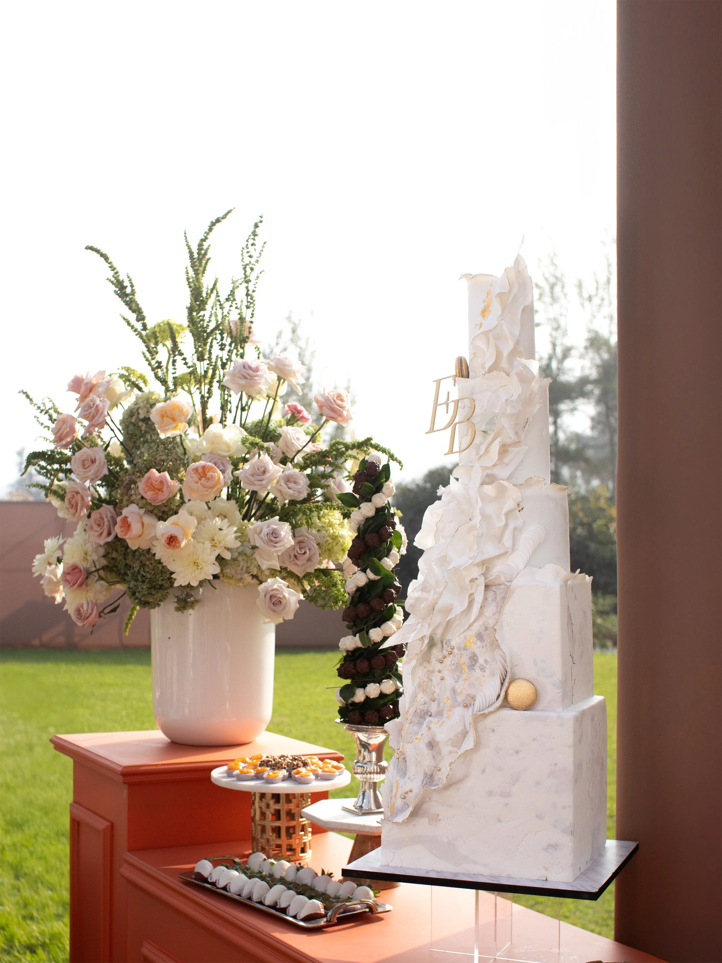 Elegant white three-tiered wedding cake with artistic decoration and gold accents, placed on a black base, next to a large floral arrangement of pink and white flowers in a white vase, with a tray of assorted small desserts and chocolates on a coral-