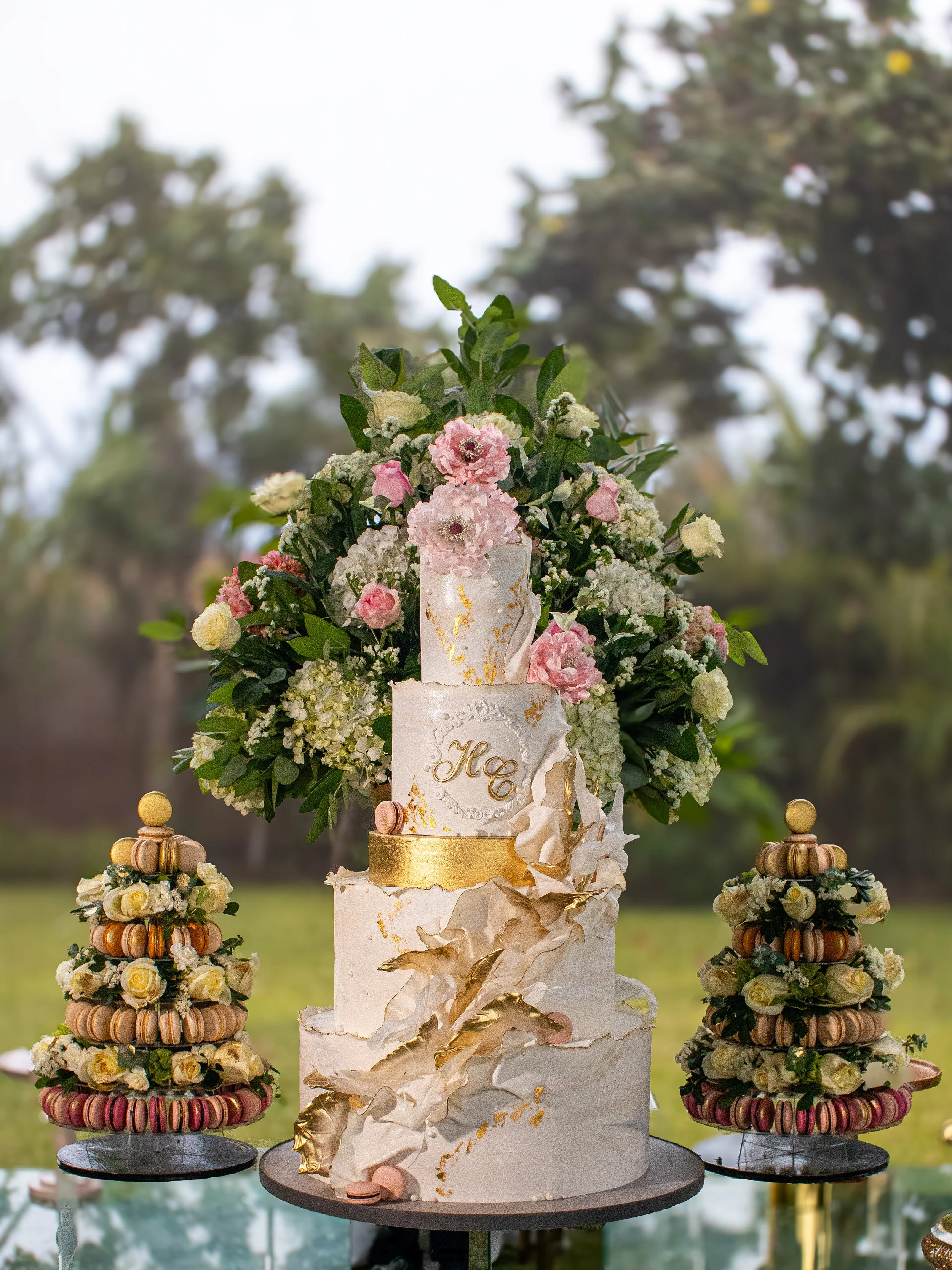 Elegant wedding cake with floral decorations and gold accents, surrounded by macarons, outdoors with trees in the background.