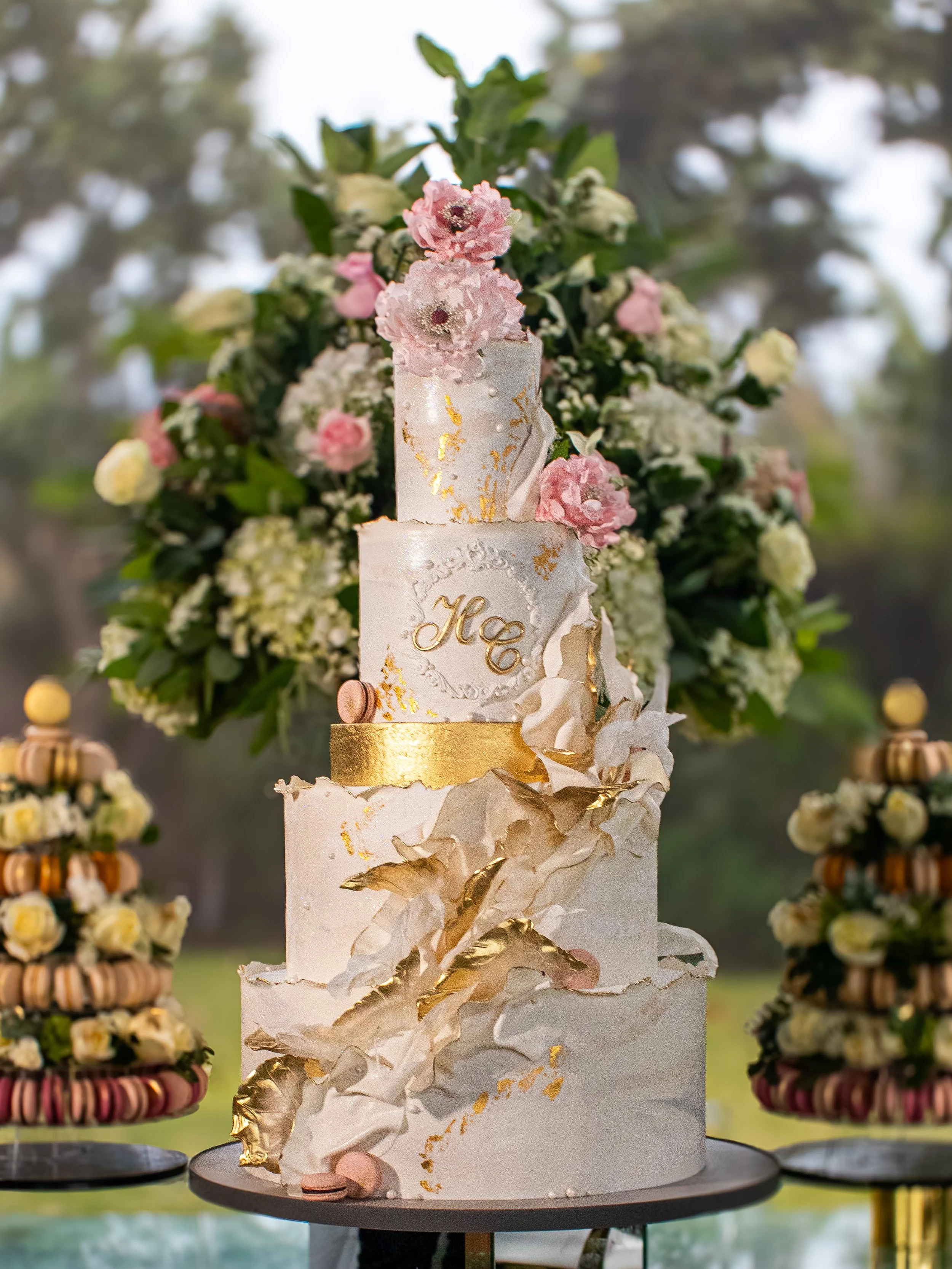 A tall, elegant white wedding cake decorated with gold accents, pink and white flowers, and gold leaf details, placed on a black cake stand. There are additional pastry towers with macarons on either side of the cake.