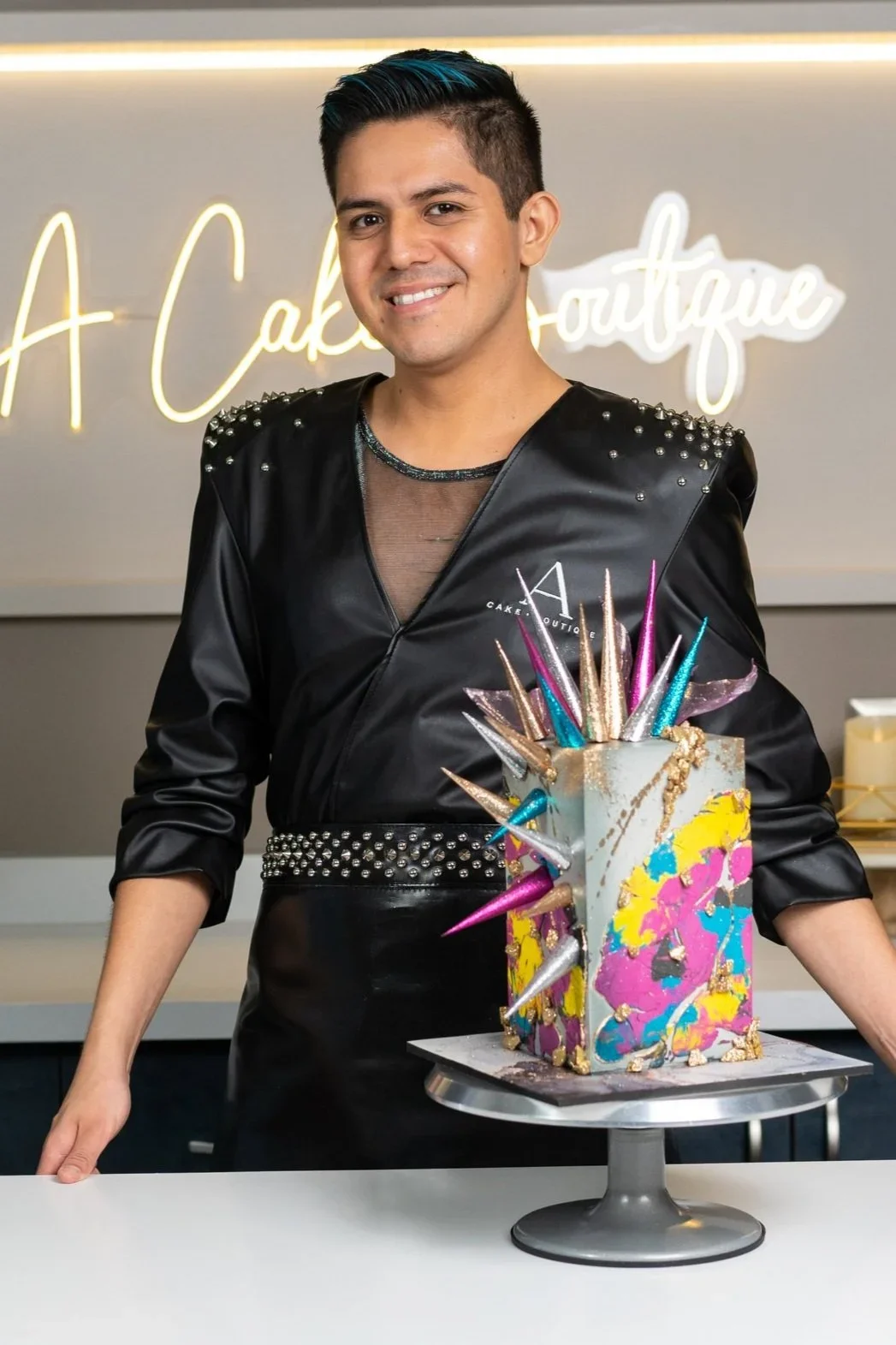 Person standing behind a colorful, modern cake with spikes on a cake stand in a bakery.
