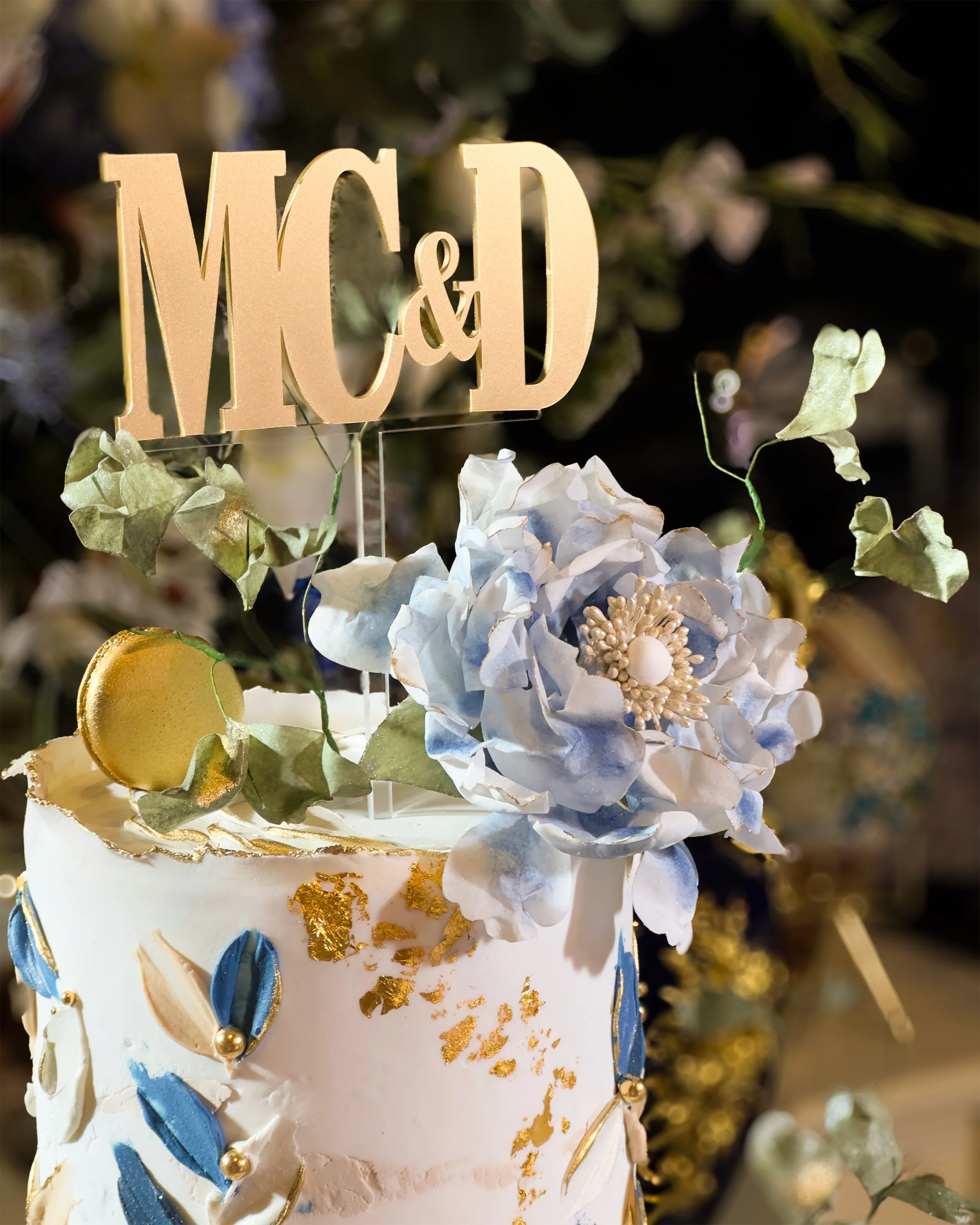 A white wedding cake decorated with blue and gold accents, a large blue flower, and green leaves, with a gold macaron on top. A wooden cake topper with the initials 'MCD' is placed above the cake.