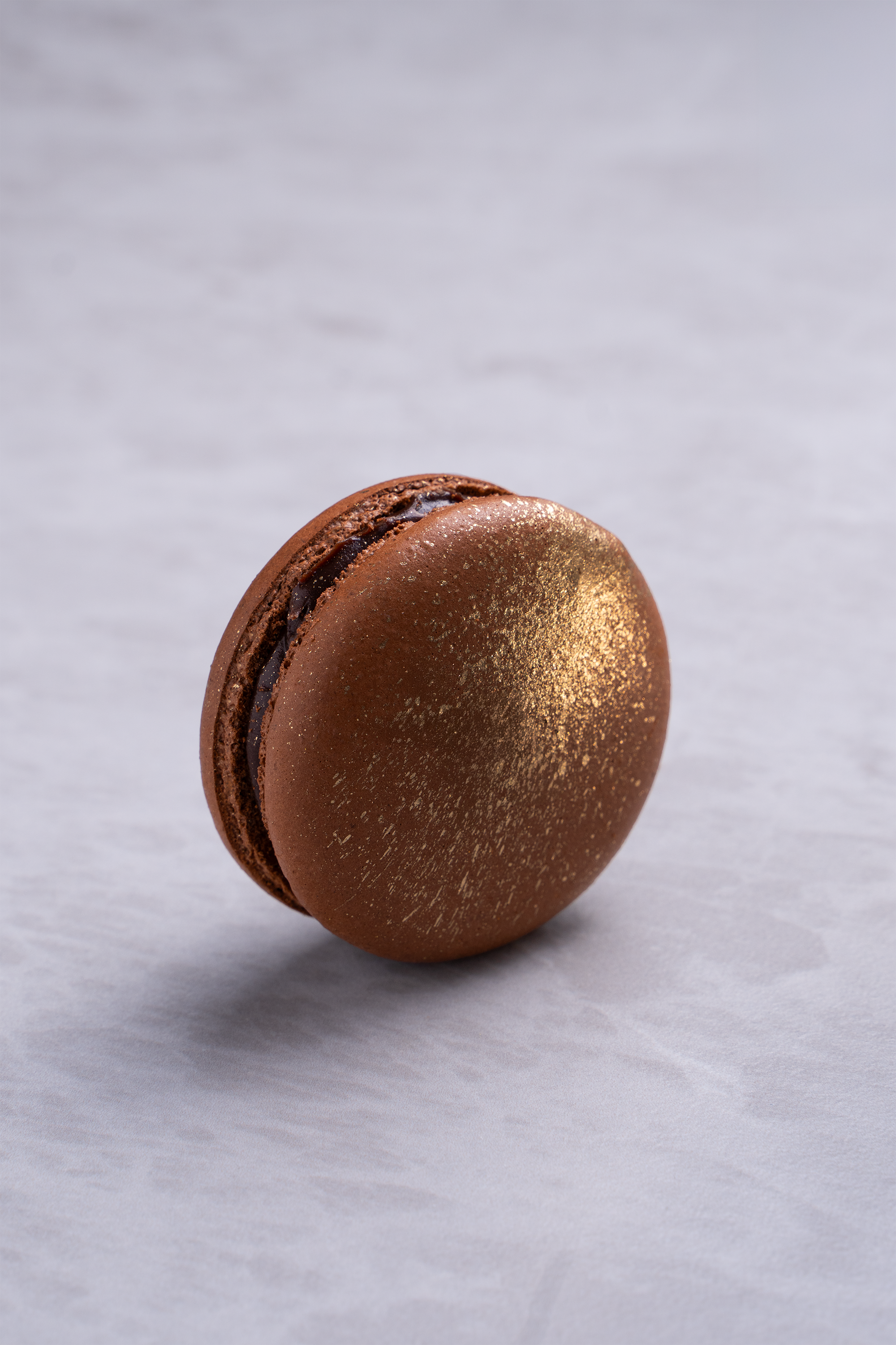 A chocolate macaron with a gold shimmer on top, placed on a light gray surface.