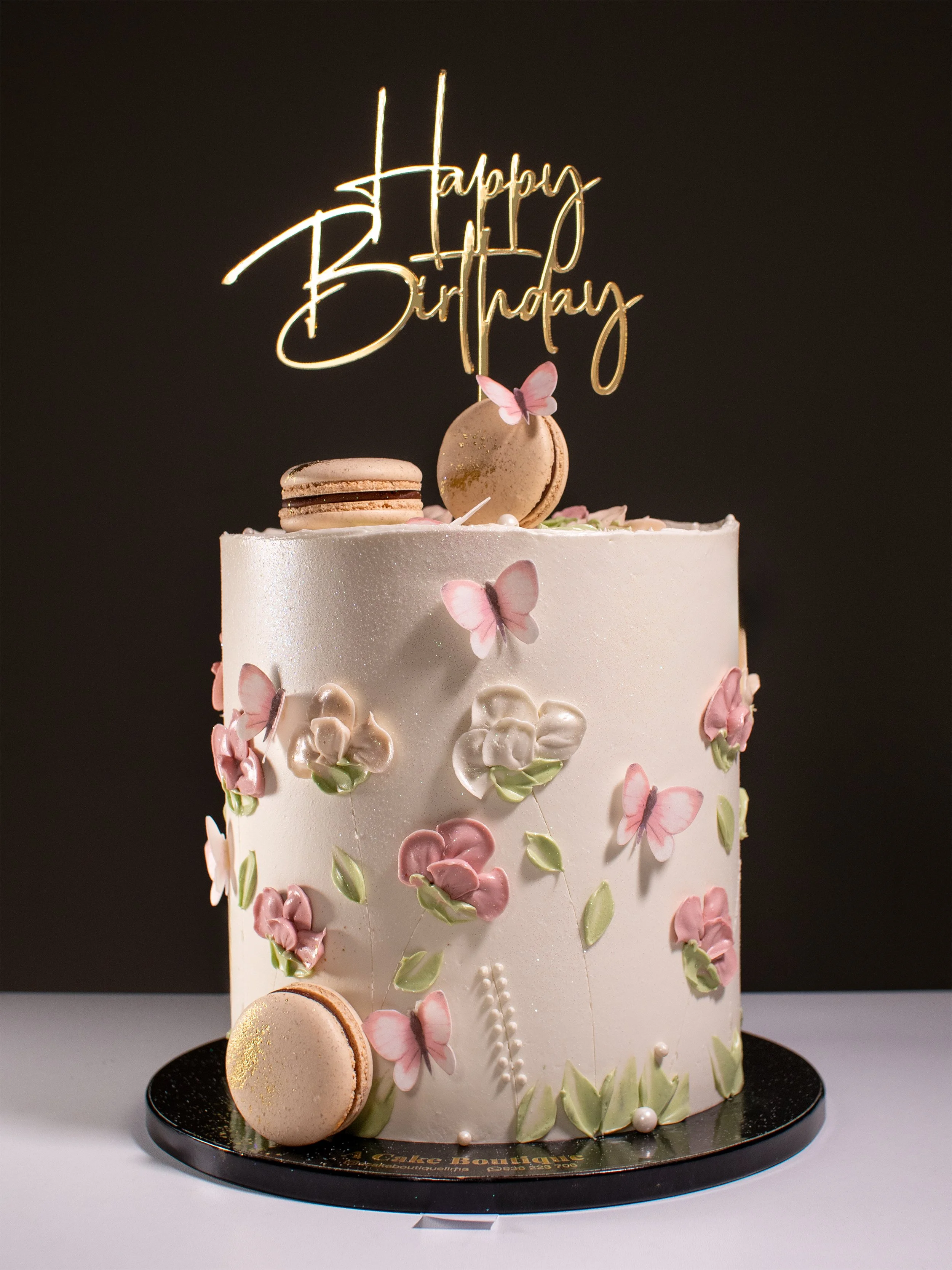 A white birthday cake decorated with pink and white flowers, butterflies, and macarons, topped with a gold "Happy Birthday" topper, on a black round base against a black background.