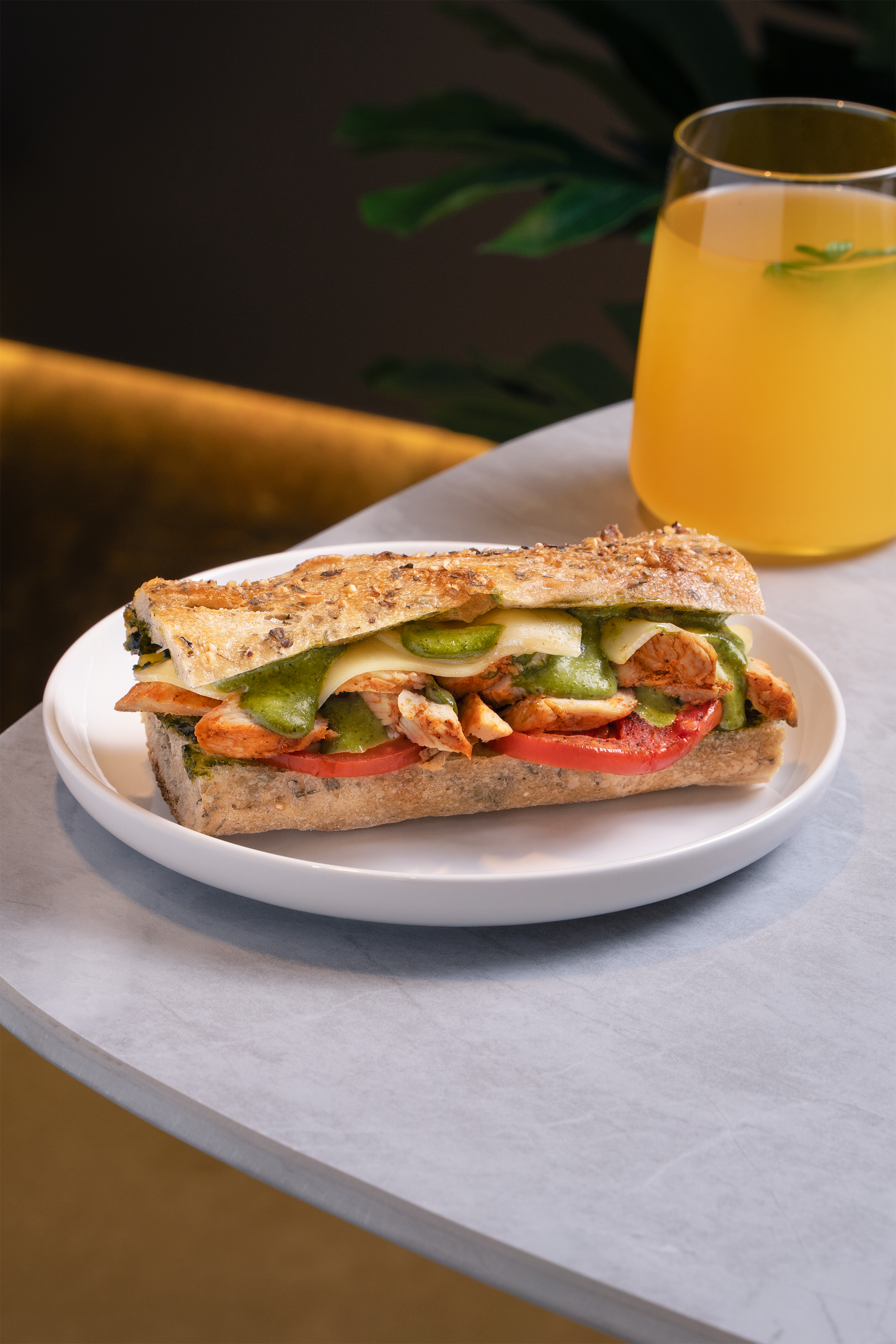 A sandwich with grilled bread, turkey, tomato, green pepper, cheese, and other vegetables served on a white plate, with a glass of yellow juice in the background on a gray marble table.