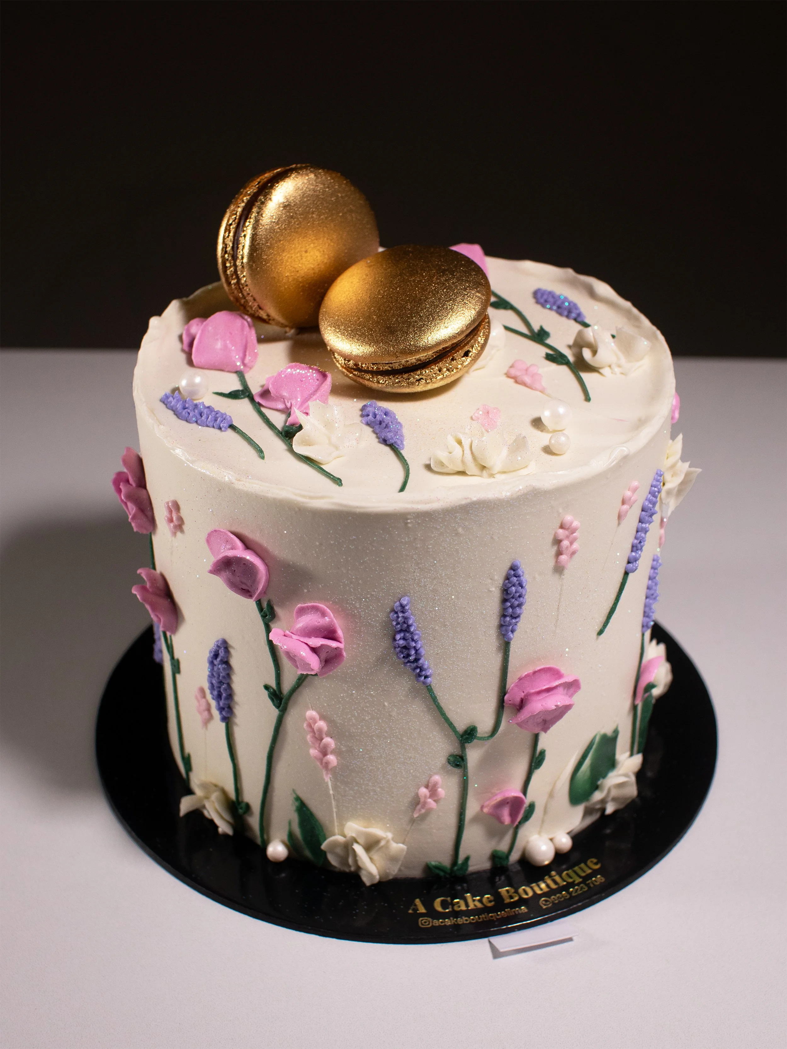 A decorated cake with white icing, pink and purple floral designs, topped with two gold macarons and small decorative pearls.