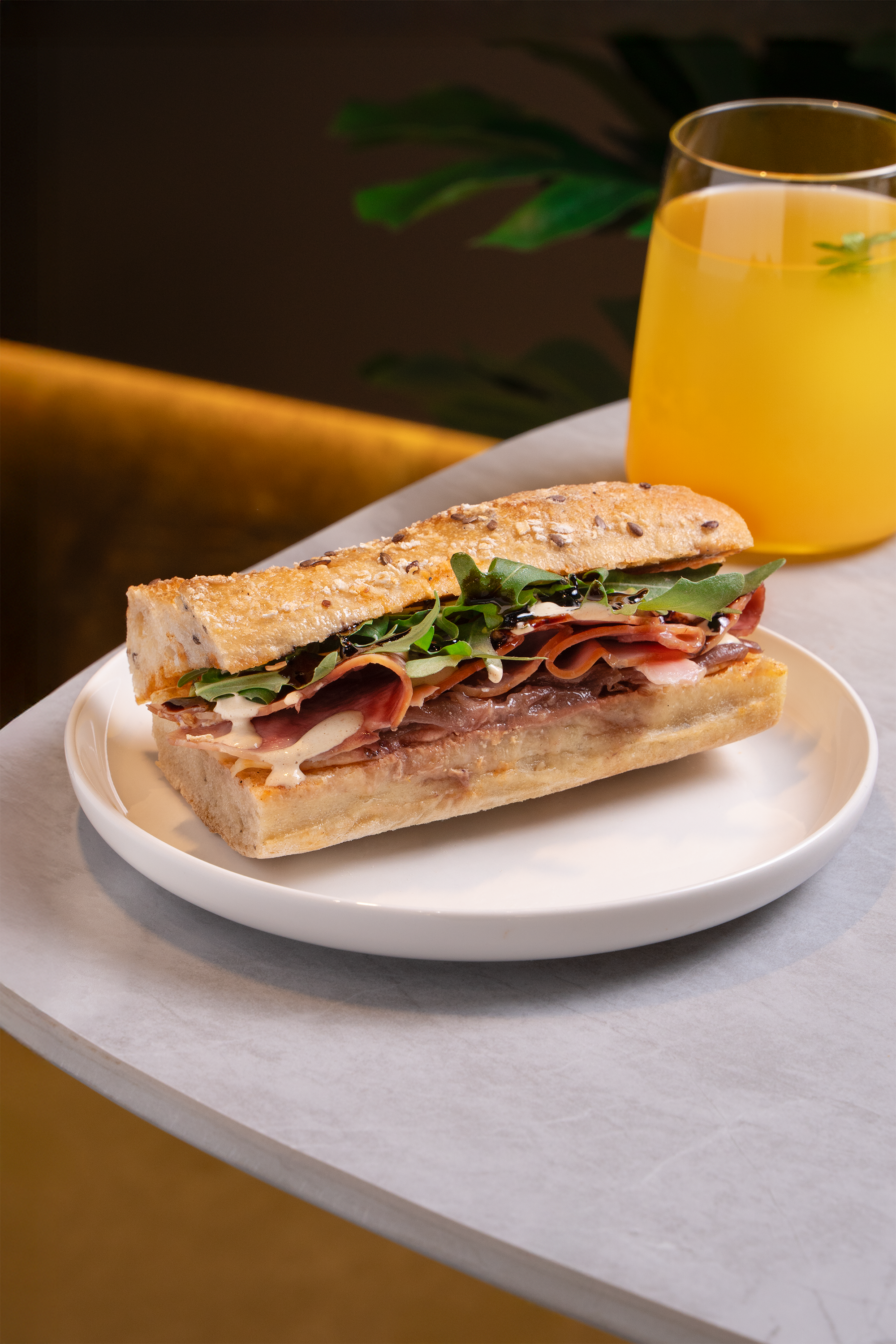 A sandwich with thinly sliced meat, leafy greens, and sauce in a baguette, served on a white plate, with a glass of orange juice in the background.