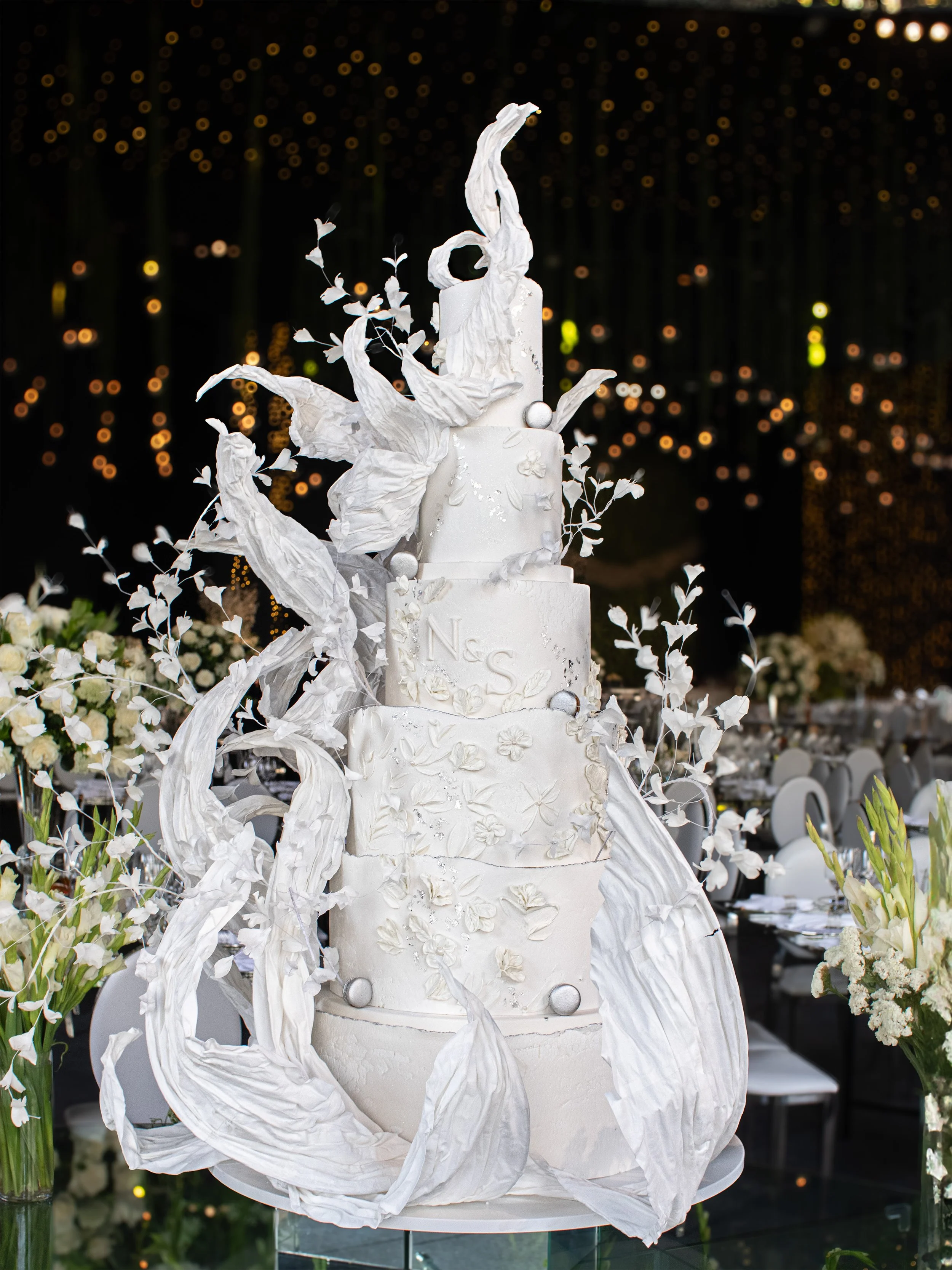A tall, elegant white wedding cake decorated with white flowers, large white leaves, and silver spherical accents, set in a decorated reception hall with floral arrangements and string lights.