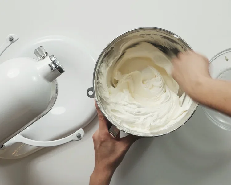 Person mixing creamy batter in a metal bowl with a hand mixer, on a white countertop.