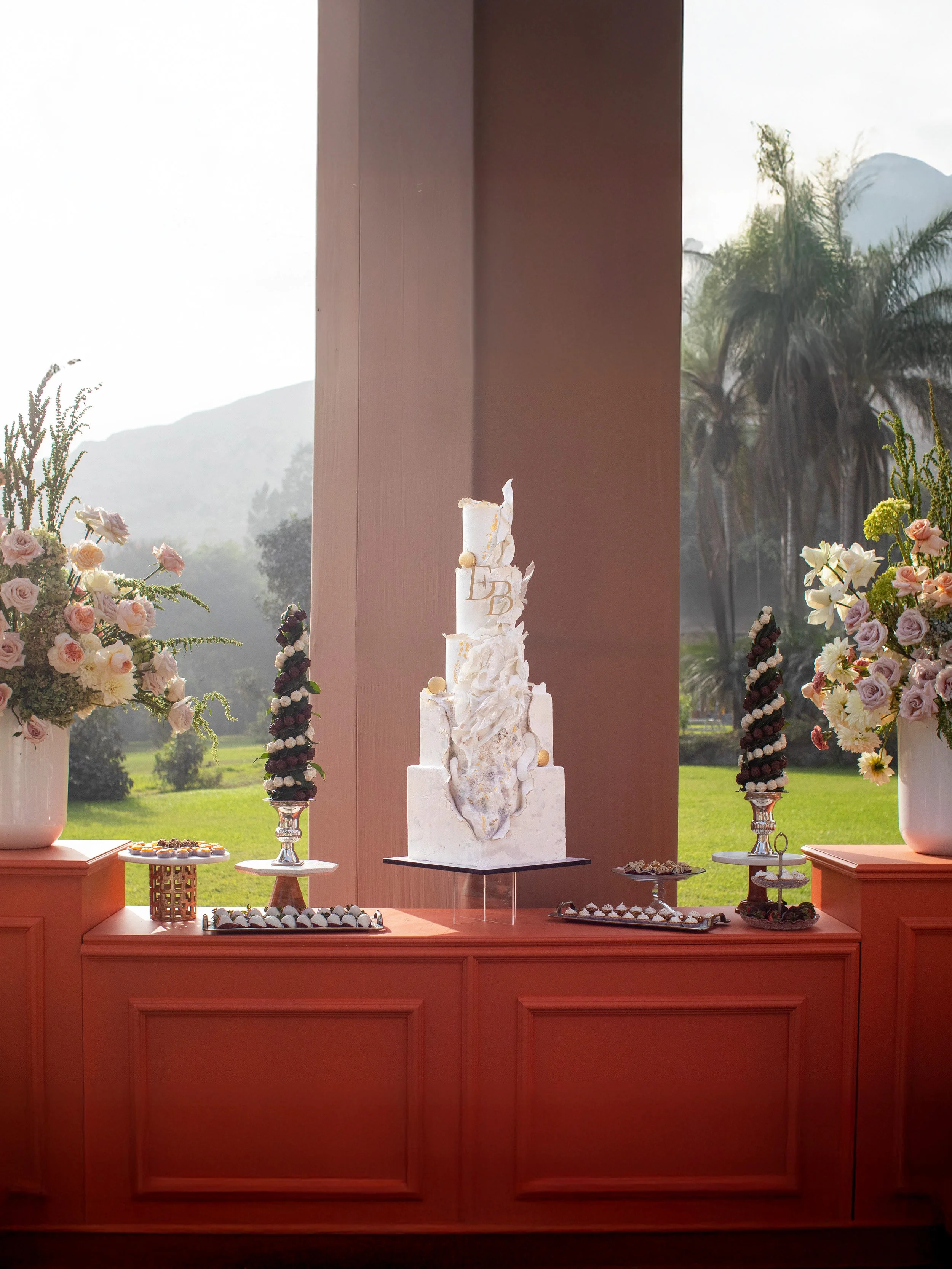 Elegant white tiered wedding cake with decorative white flowers, displayed on a stand in front of a large window with a scenic outdoor view of greenery and mountains.