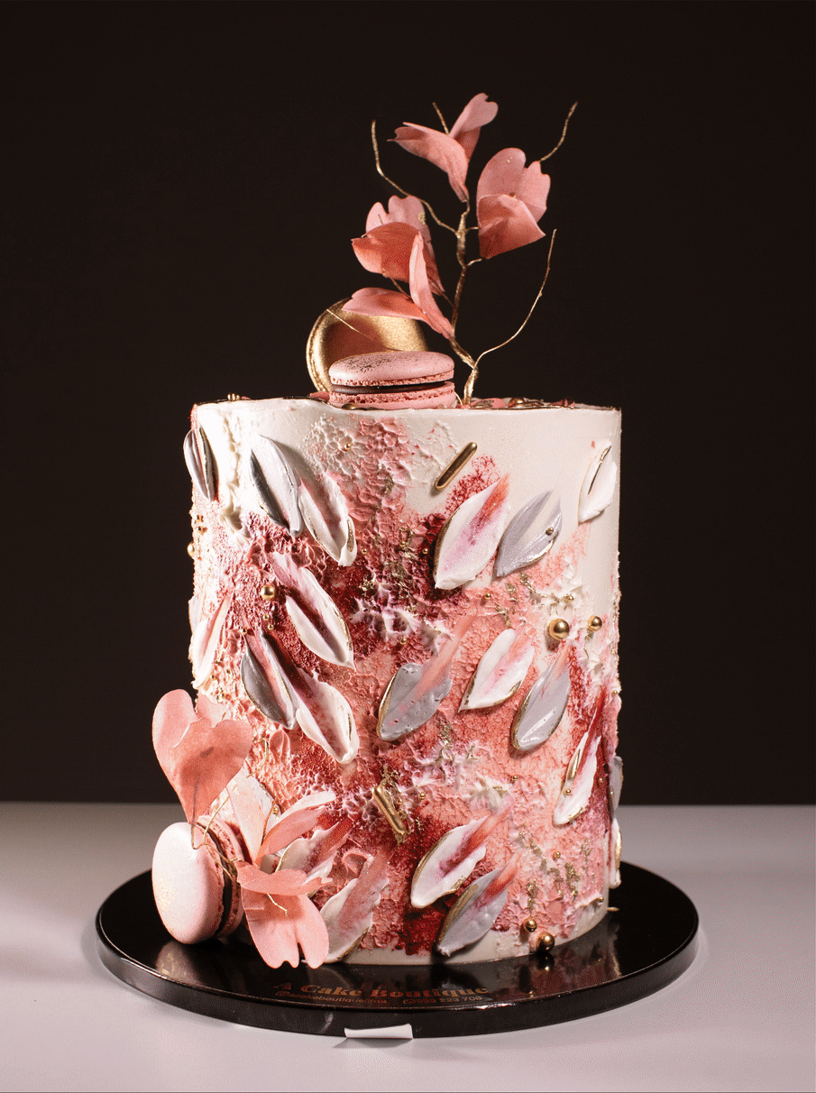 Elegant pink and white decorated cake with floral and metallic accents, topped with a macaron and pink flowers, placed on a black cake board against a black background.