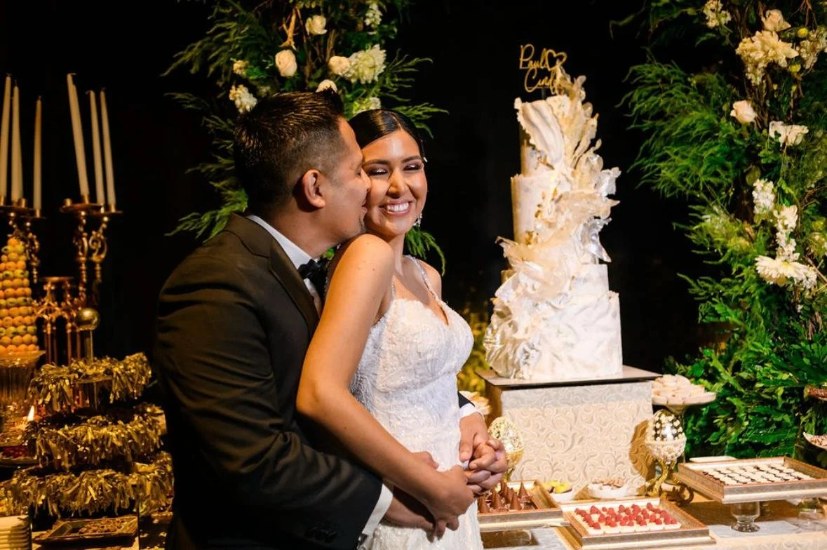 A bride and groom sharing a special moment at their wedding, with the groom whispering into the bride's ear as she smiles. The background features a tiered wedding cake and elegant decorations with green foliage and white flowers.