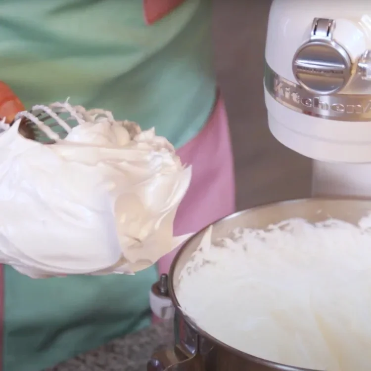 Person holding a bowl of whipped cream or frosting next to a stand mixer with more whipped cream or frosting inside.