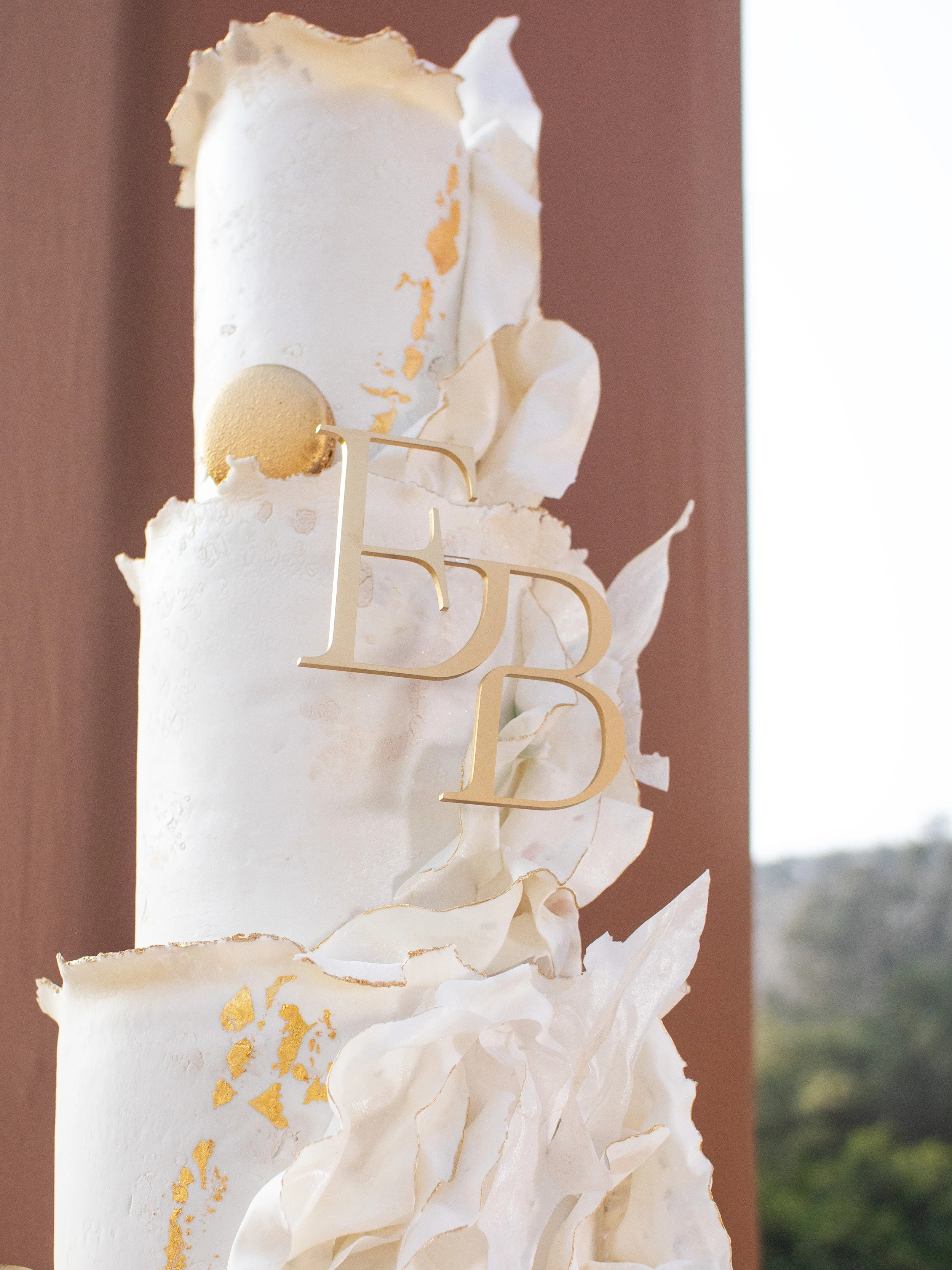 A tall white wedding cake with gold accents and decorations, topped with a gold-colored ball, and a gold "EDD" cake topper.