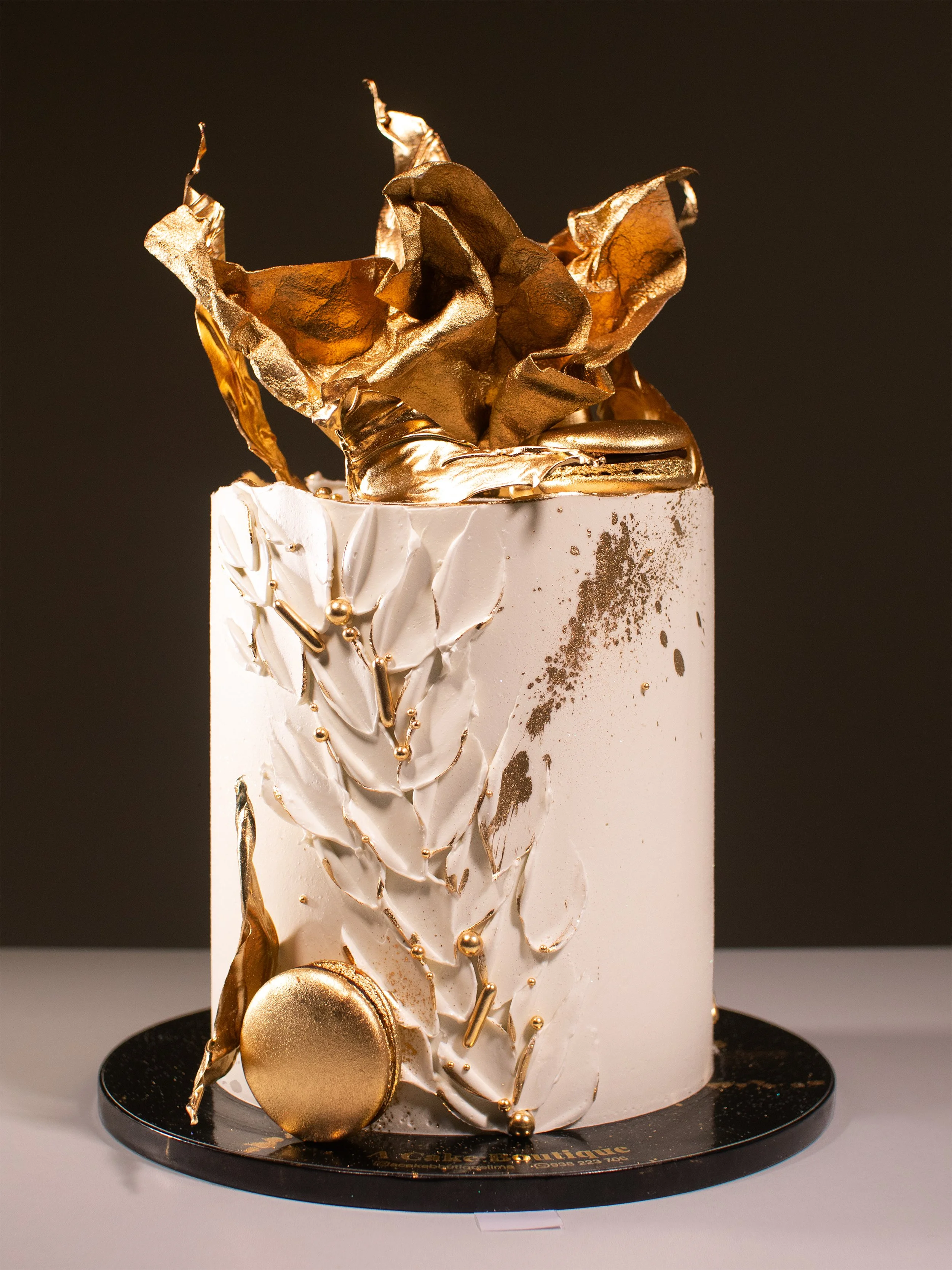 A tall, cylindrical cake with white icing decorated with gold-colored geometric and organic shapes, including leaves and spheres, with a dark background.