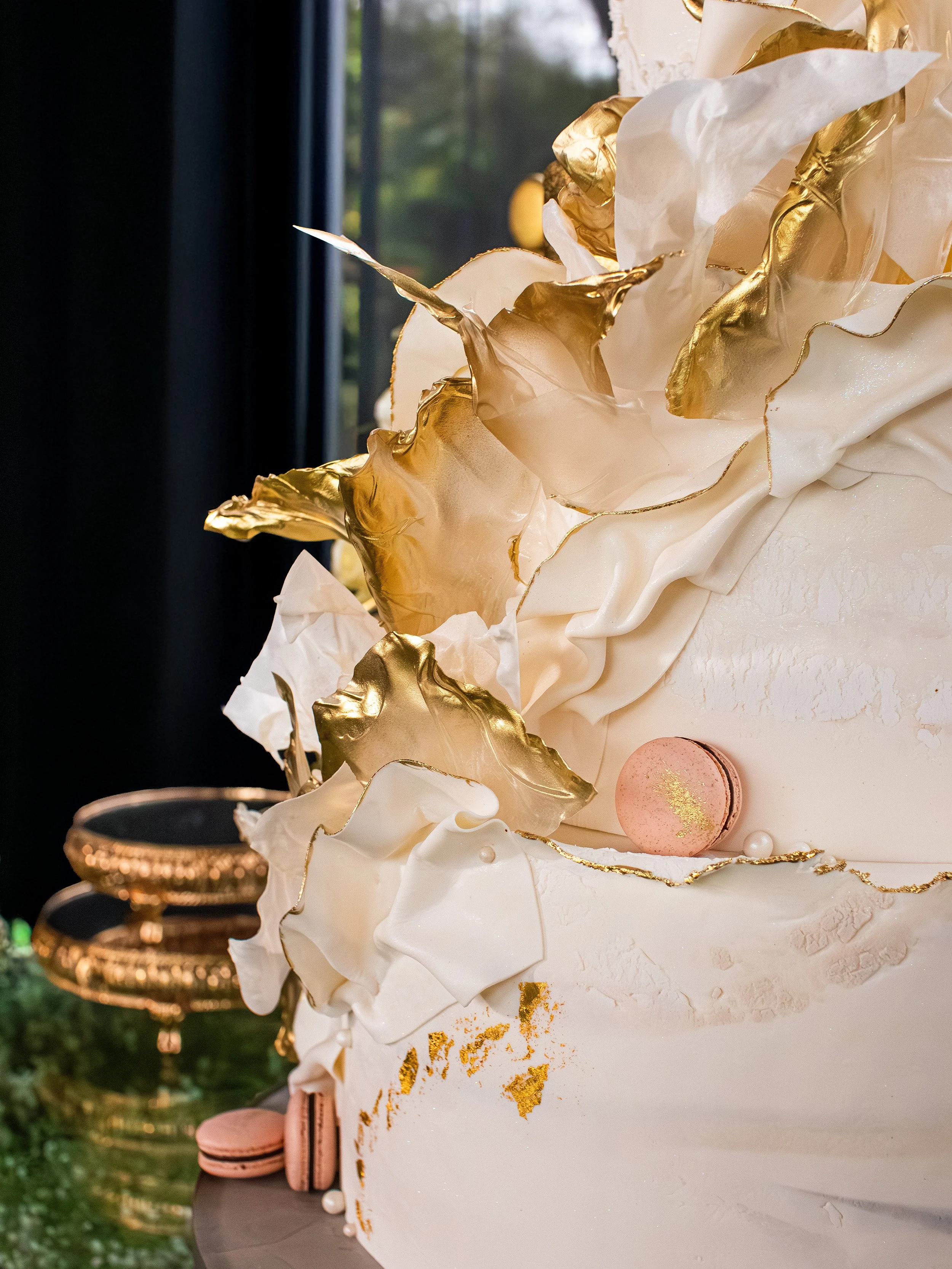 Close-up of a decorated cake with white icing, gold accents, and pink macarons, with gold and white leaf-like decorations on top, set against a blurred background with a dark curtain and trees outside window.