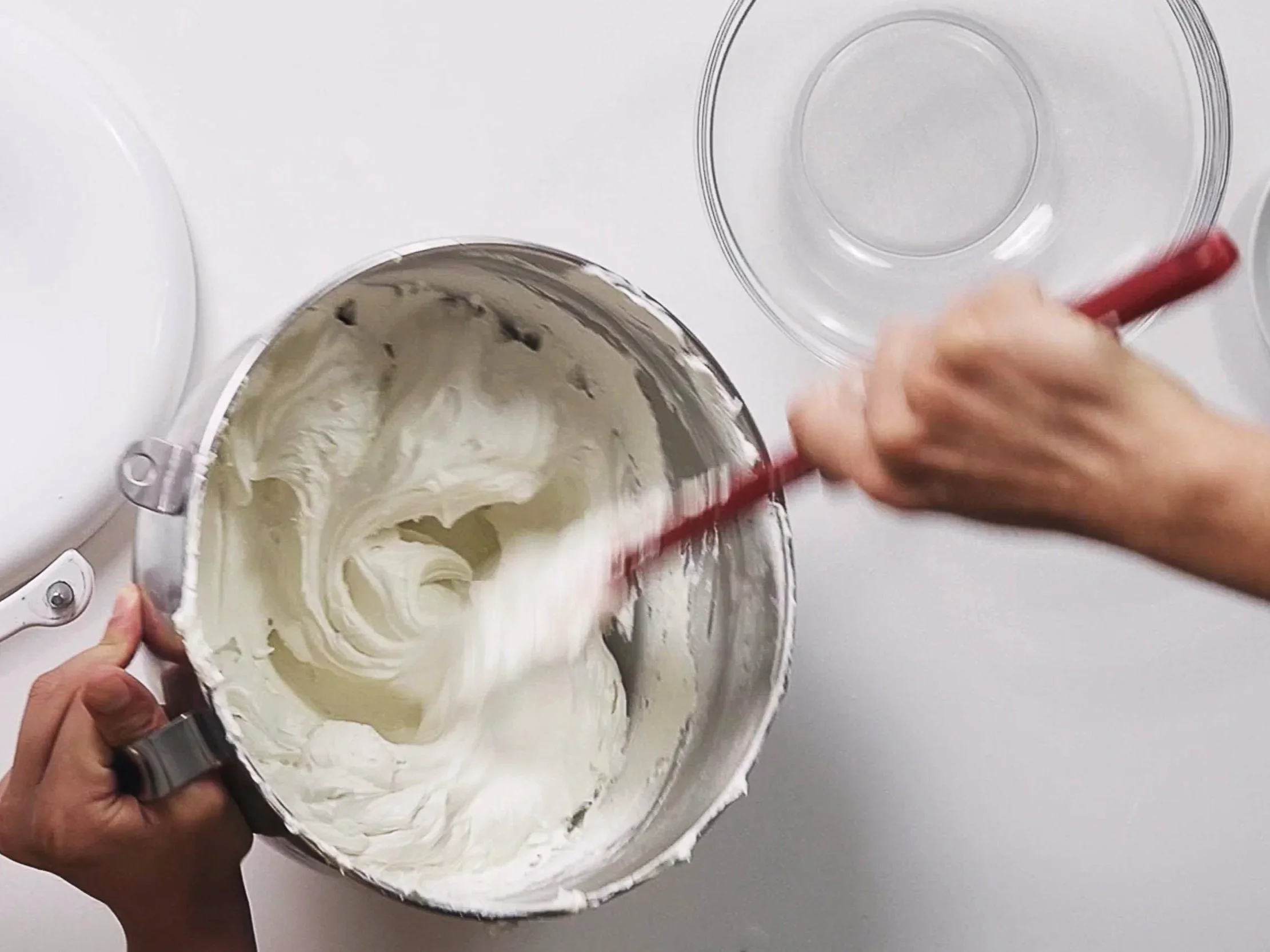 Person mixing whipped cream or frosting in a metal bowl with a red spatula on a white surface. Clear glass bowls are nearby.