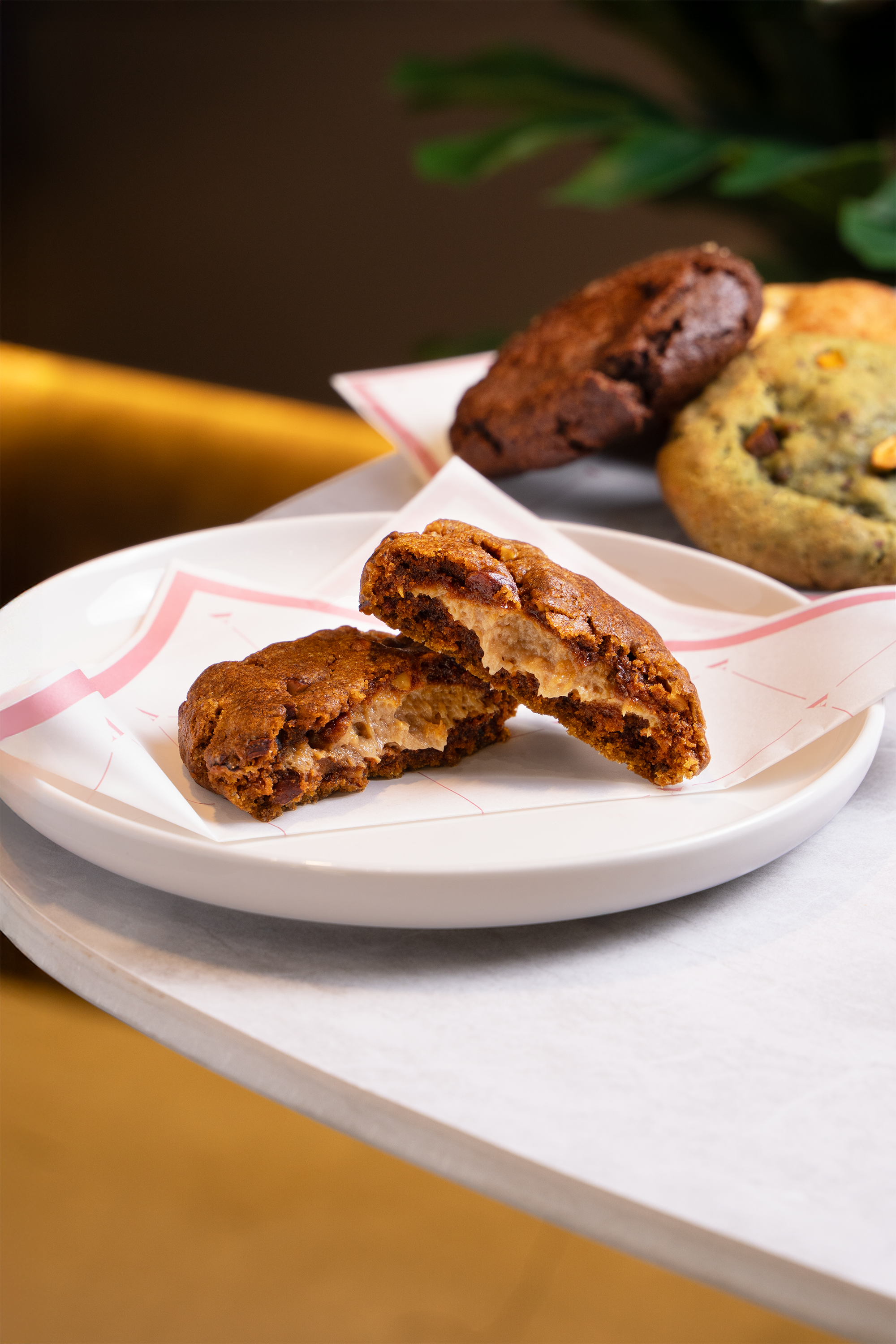 Close-up of a plate with broken chocolate chip cookie, revealing its interior, placed on a white paper-lined plate on a marble surface with other cookies in the background.